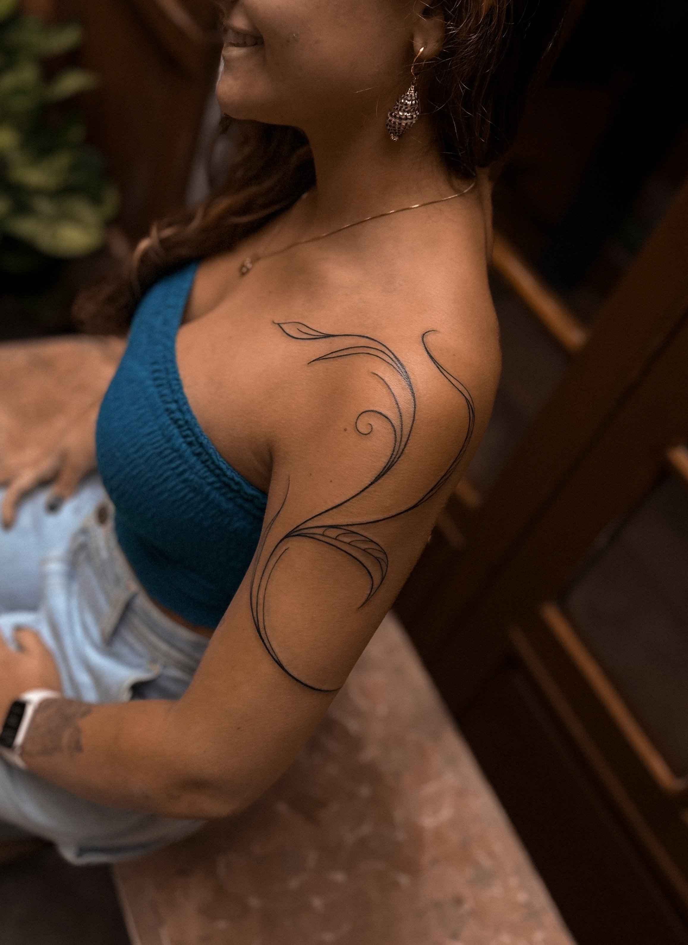 A woman with long dark hair wearing earrings, a necklace, a blue crop top, and light-colored shorts, showing a delicate blackline floral tattoo on her upper arm near her shoulder.