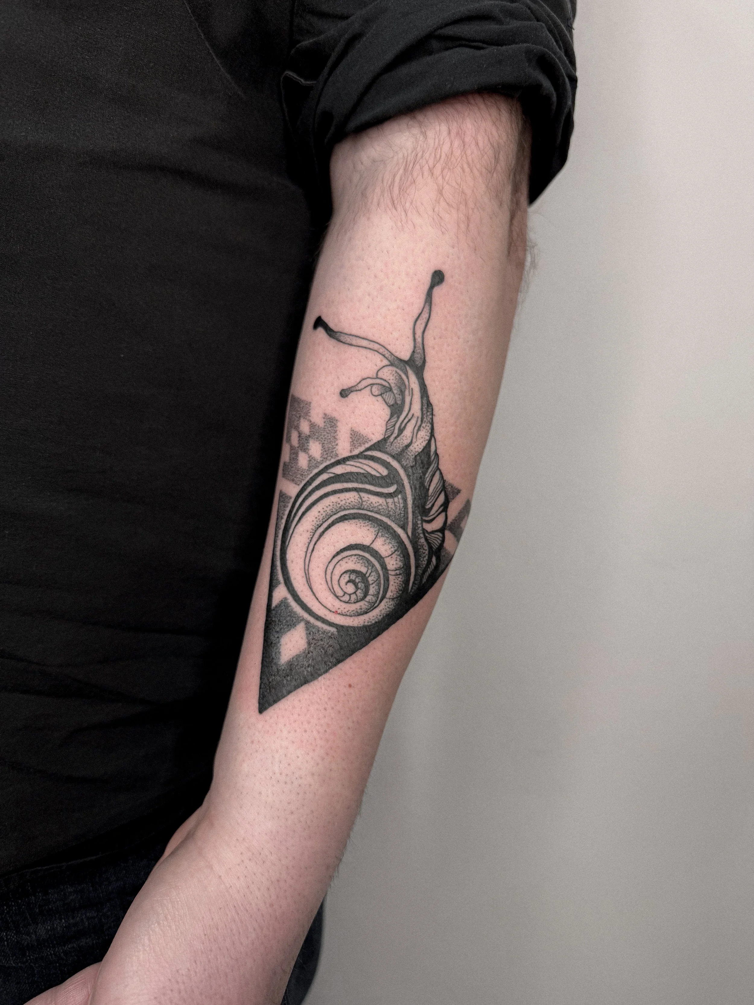 Black ink tattoo of a snail with a coiled shell on inner forearm, with geometric patterns in the background.
