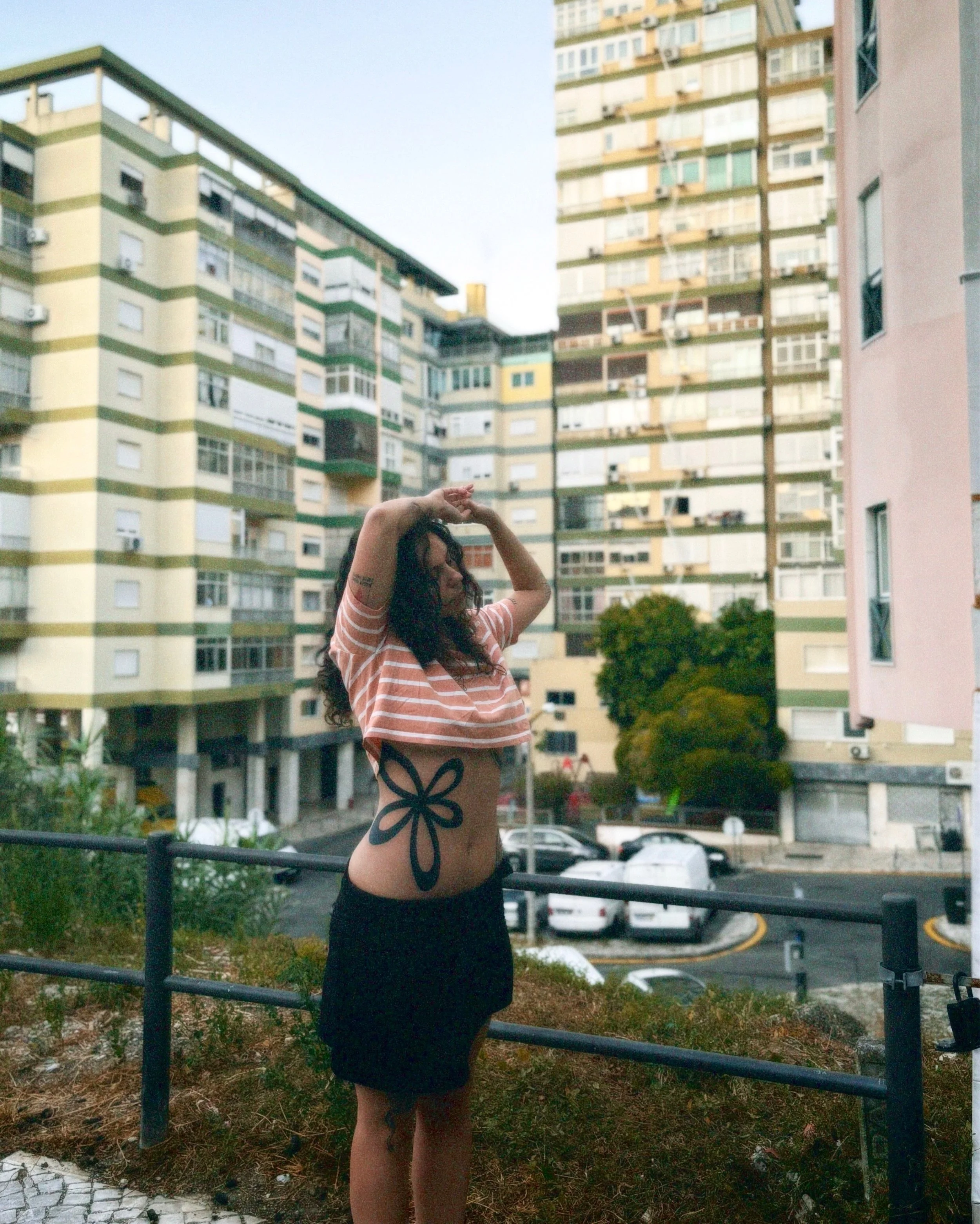 Young woman with long curly hair and tattoos, wearing a striped crop top and a black skirt, standing outdoors in front of an apartment complex, with her arms raised above her head.