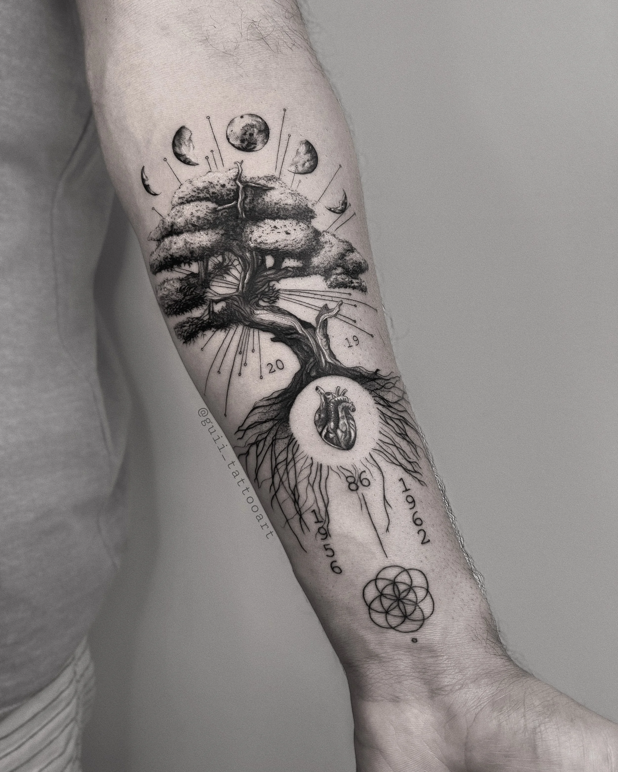 Forearm tattoo featuring a tree with a heart at the base, various phases of the moon above, numbers "1956," "1962," "1986," and "2019," and a geometric flower-like pattern near the wrist.
