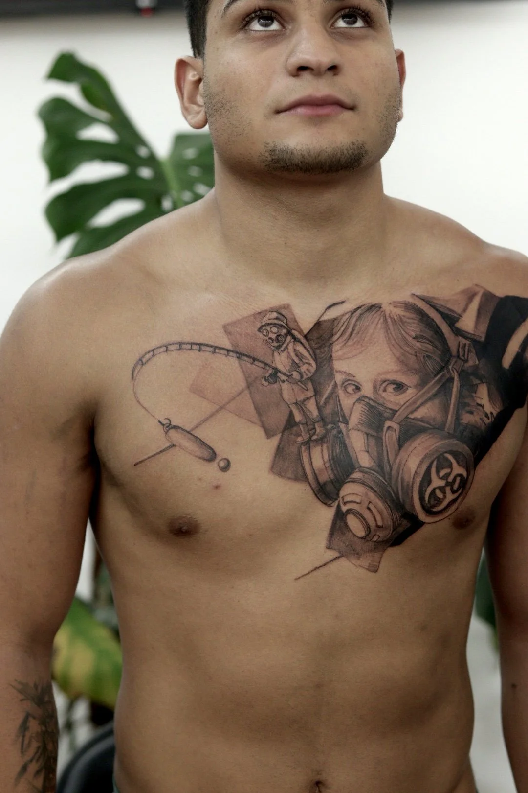 Man with detailed tattoo on chest featuring a large gas mask design and a smaller figure wearing a protective suit holding a fishing rod, with artistic elements and a leafy plant in the background.