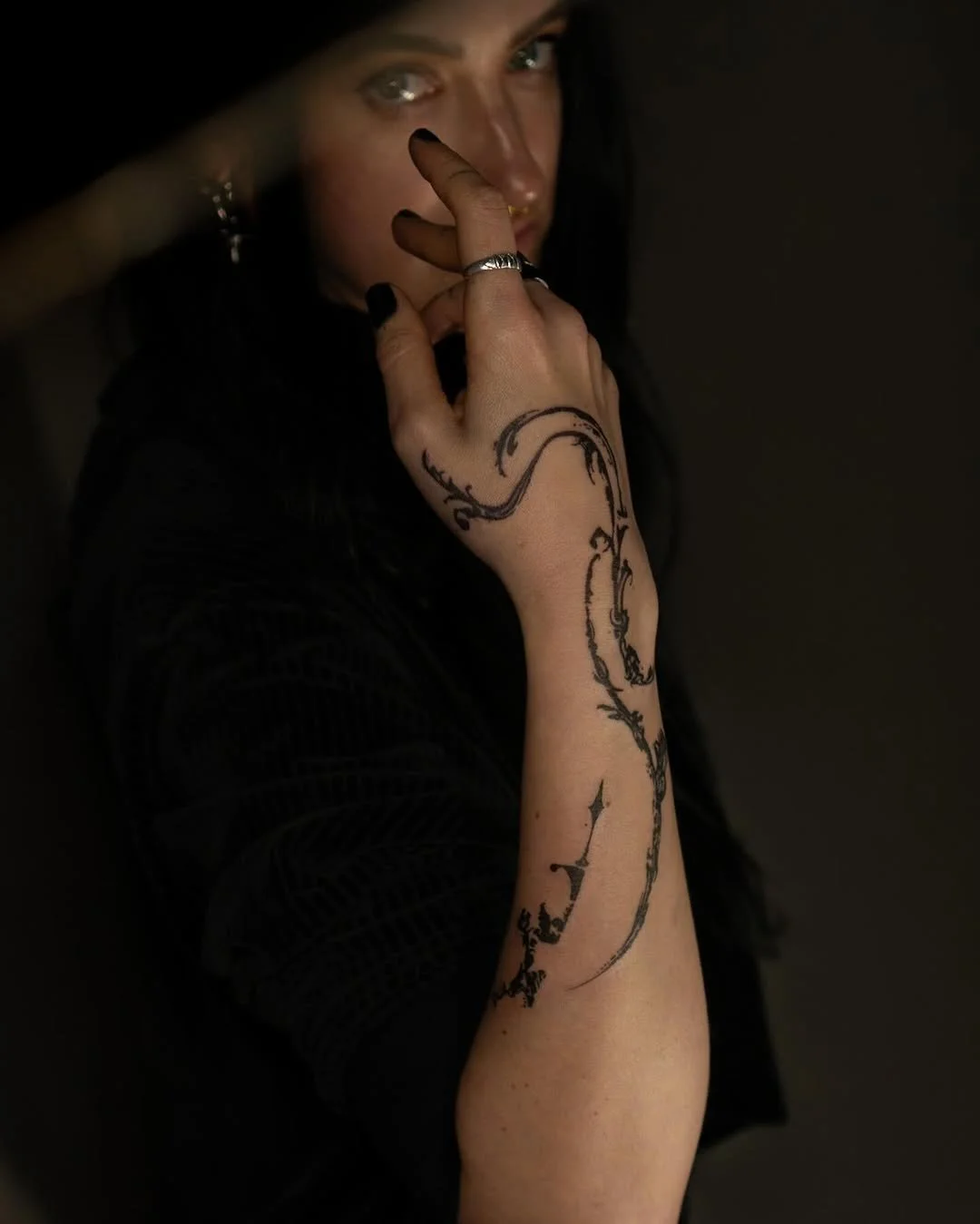 Person with a black floral tattoo on arm, wearing a ring, and dark clothing, in a dimly lit setting.