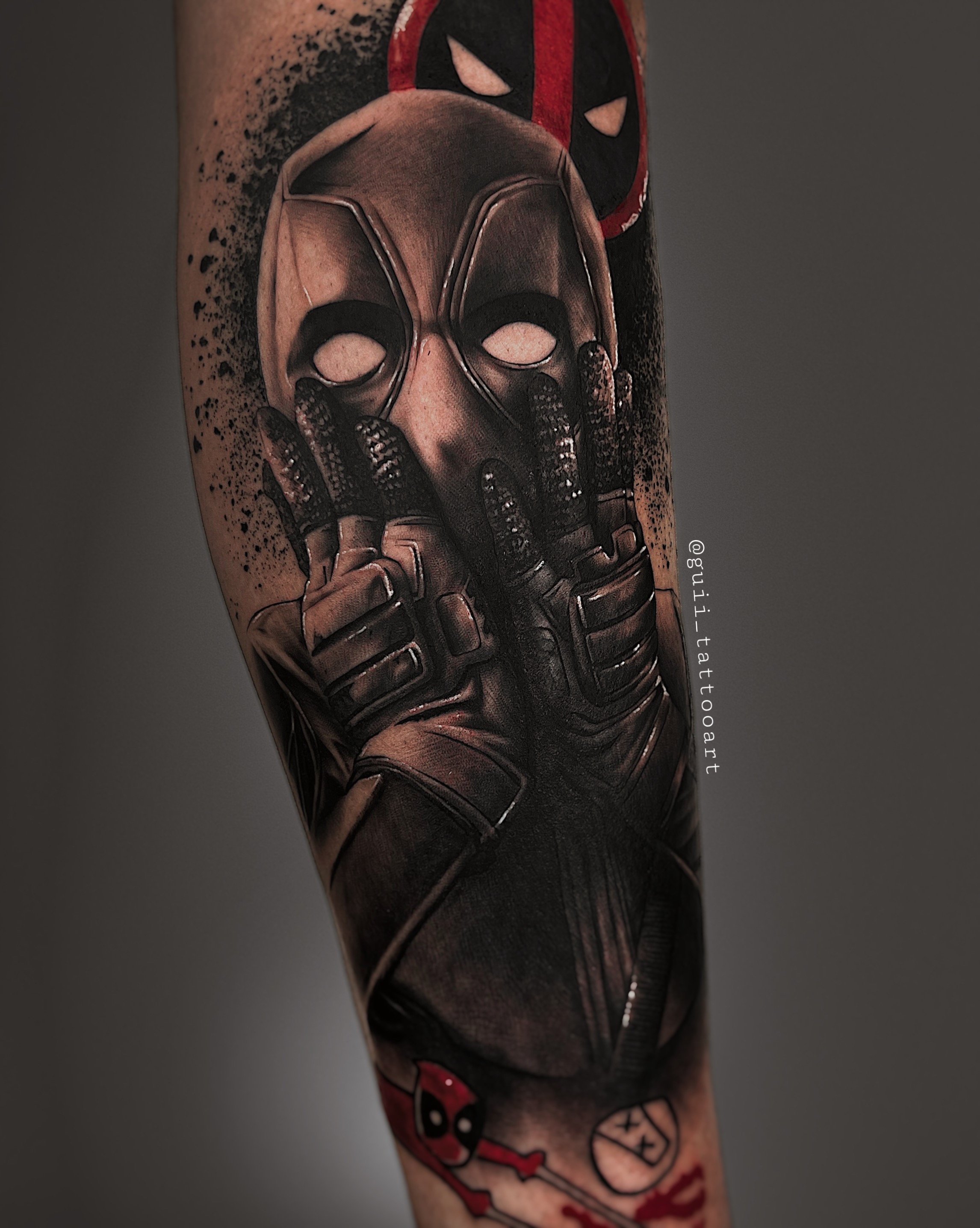 Tattoo of a comic character with hands on face, red and black mask, and a logo in the background on a person's arm.