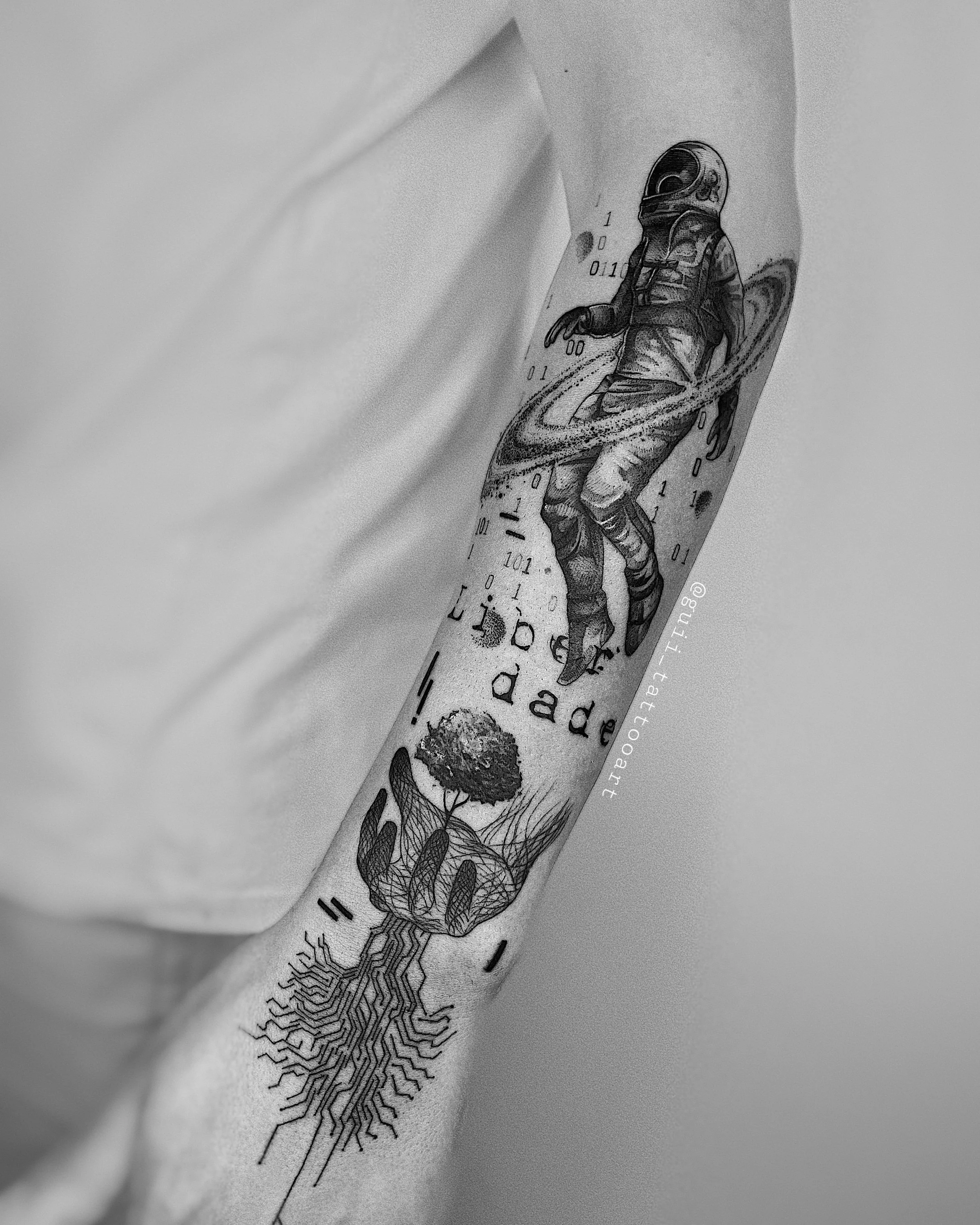Black and white tattoo on a person's arm featuring an astronaut surrounded by rings, binary code, and the words "Liberdade." Below, there is an image of a hand holding a tree with roots extending like circuits.