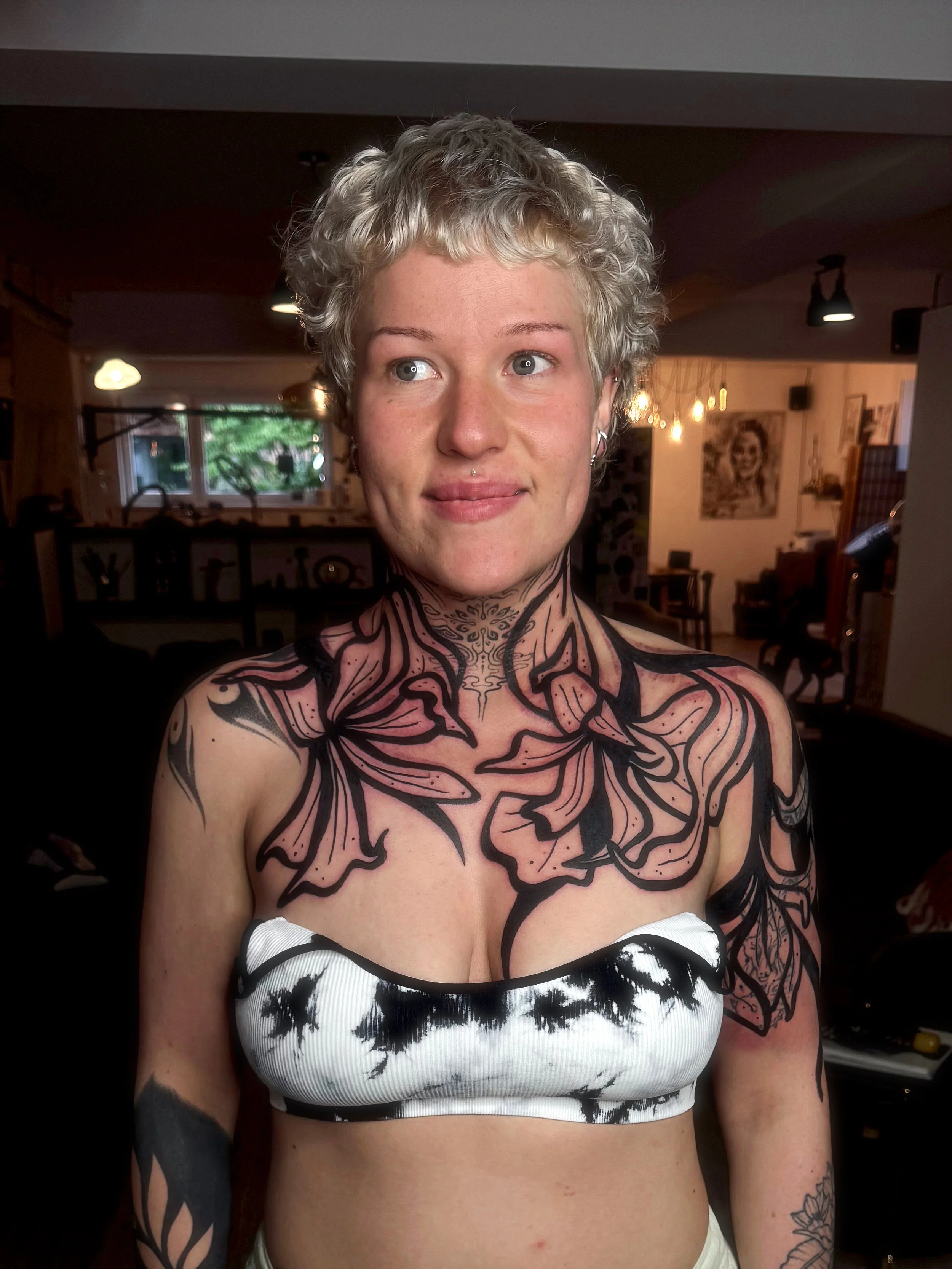 A woman with short, curly platinum blonde hair and multiple facial piercings, standing indoors with a large black and pink floral tattoo covering her chest and shoulders. She is wearing a strapless black and white top and is looking slightly to her l