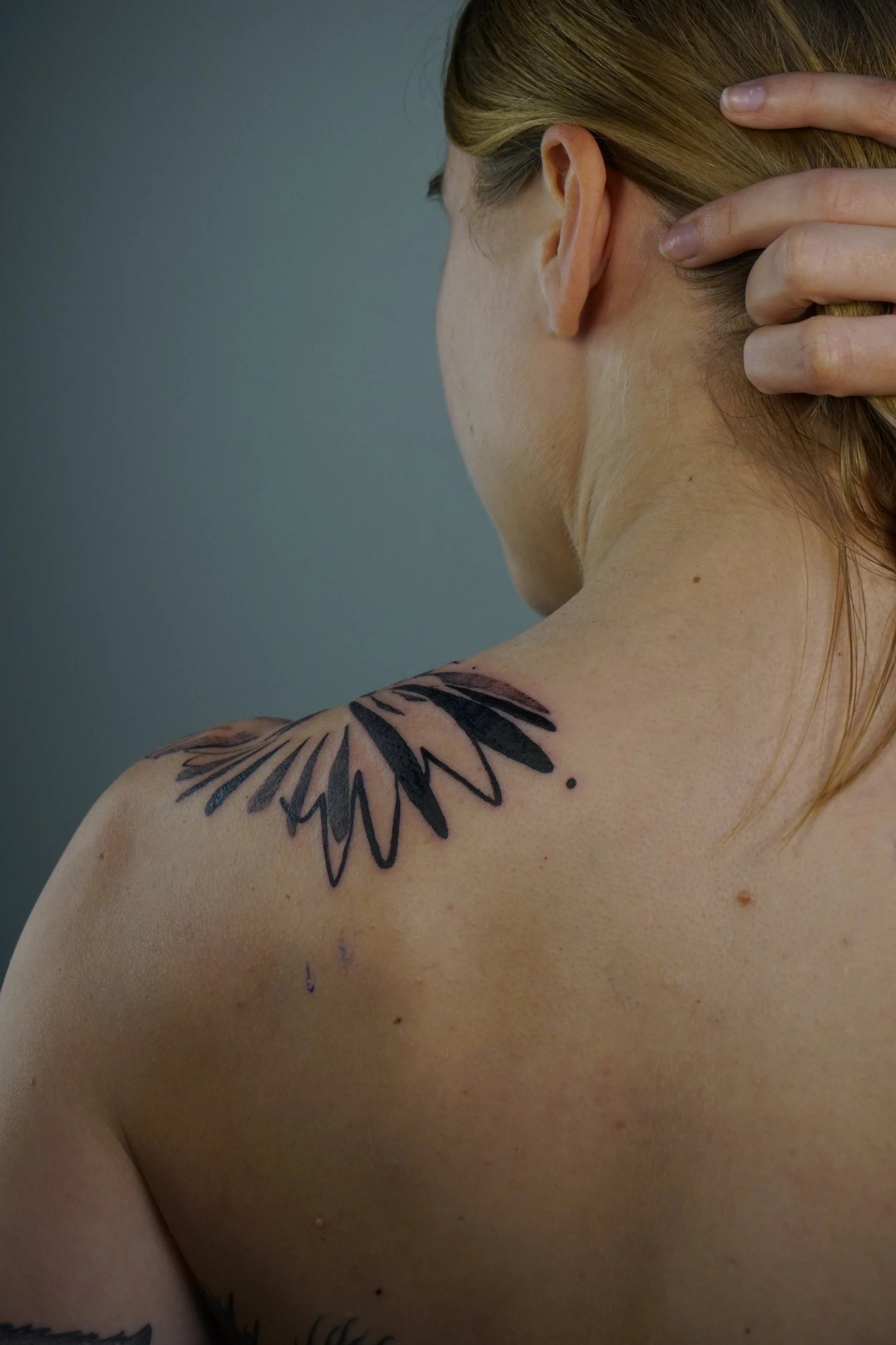 A woman with blonde hair touching her neck, showing a black tattoo of a feather on her shoulder.