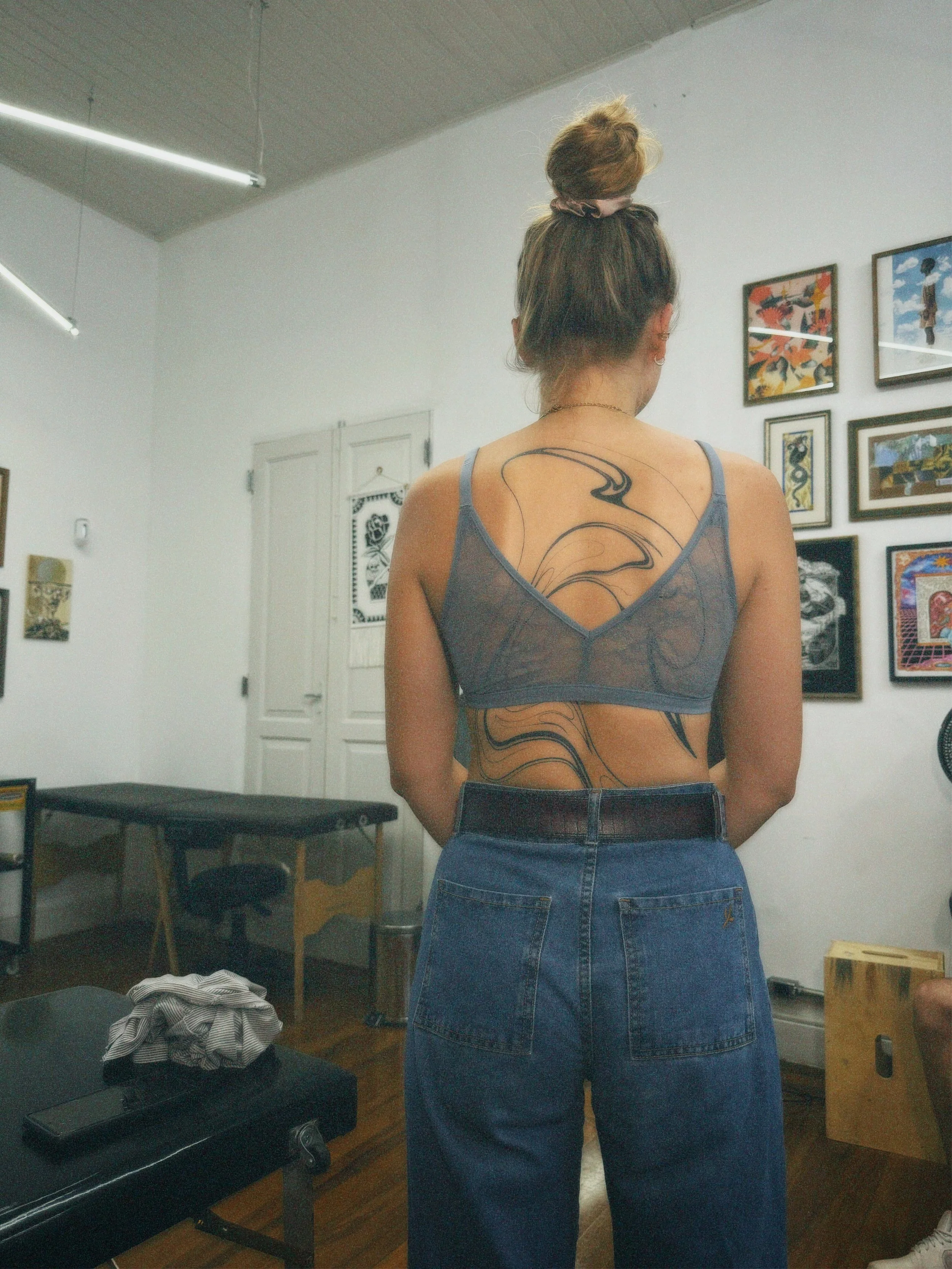 A woman with brown hair tied in a bun, wearing a semi-sheer gray camisole and high-waisted blue jeans, with visible tattoos on her back and side, stands in an art gallery looking at artwork on the wall.