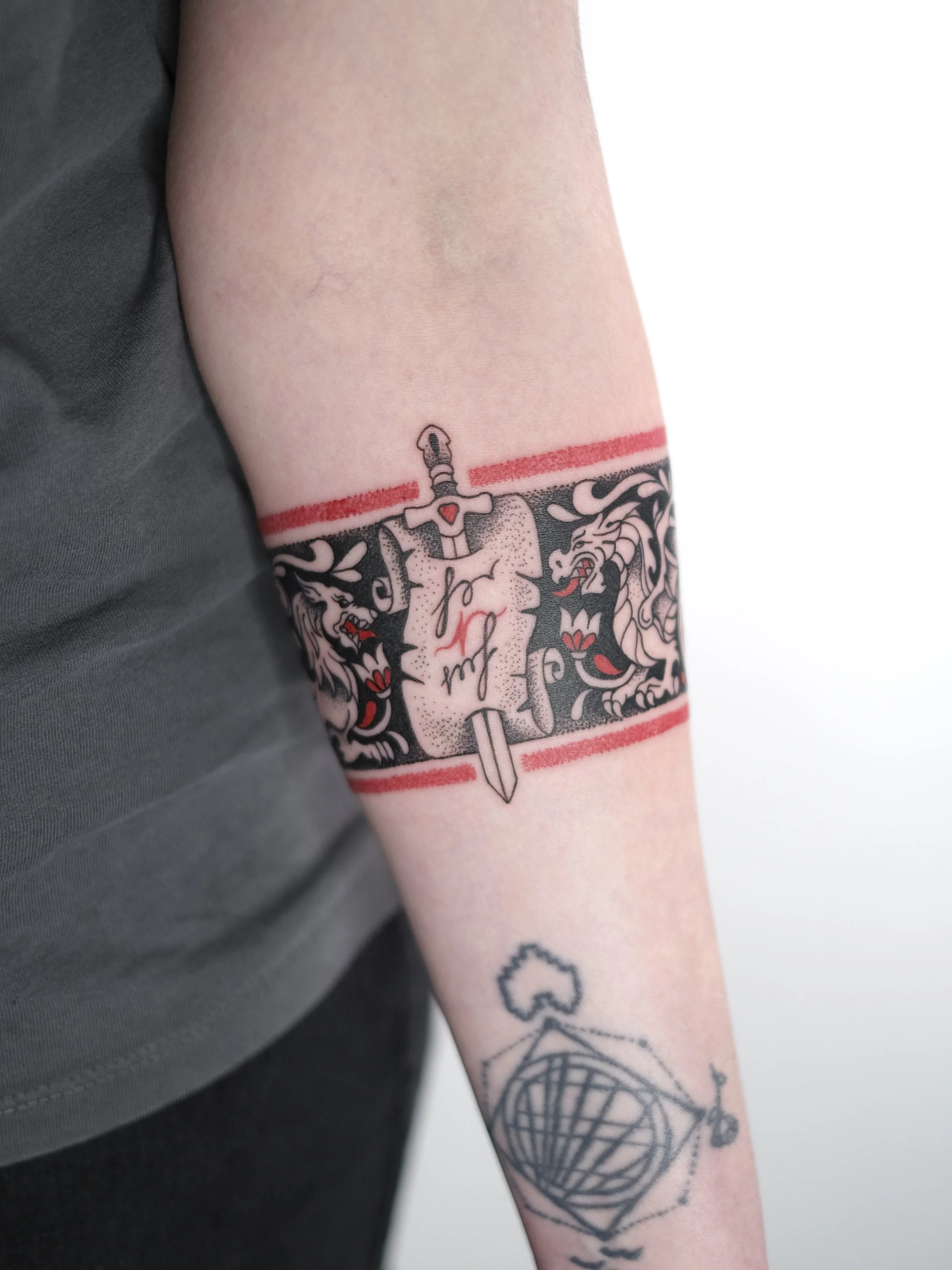 Tattoo of a sword and shield with lion heads and flames, surrounded by striped bands, on a person's upper arm.