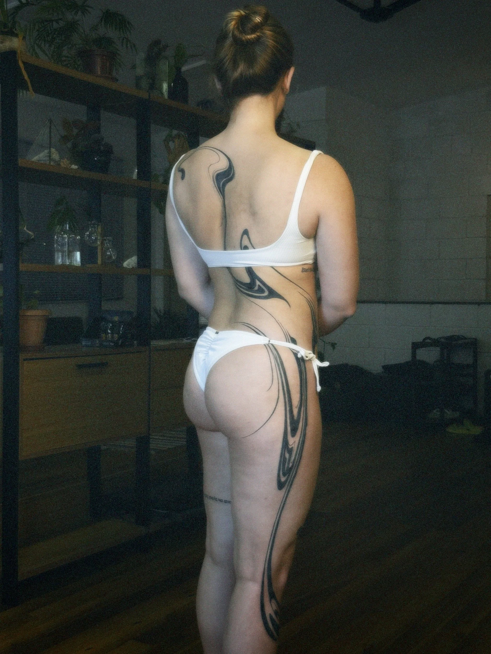 A woman with short hair styled in a bun, wearing a white bikini, shows back tattoos on her back and left leg in an indoor setting with a wooden shelf and plants in the background.