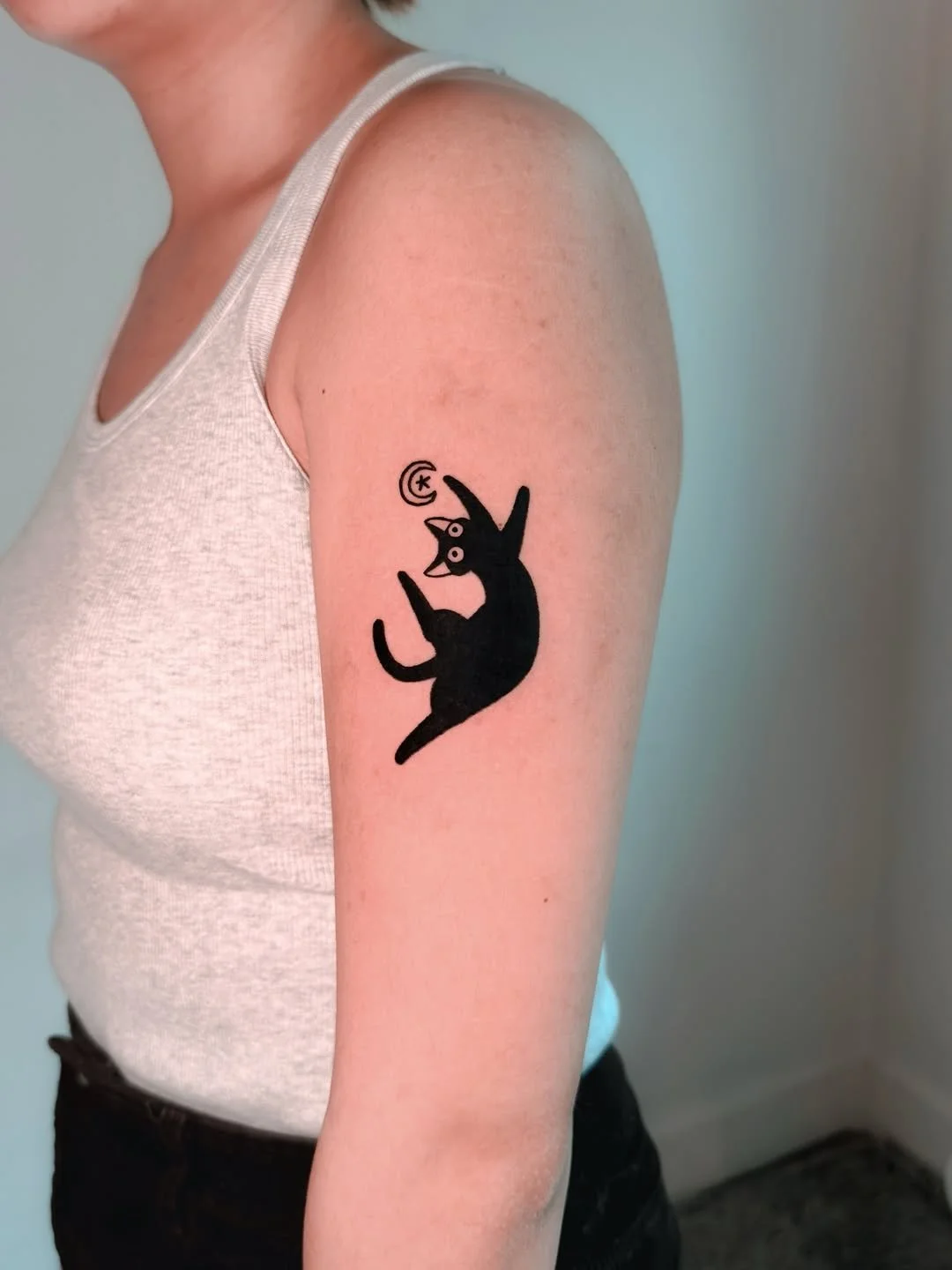 Tattoo of a black cat falling or leaping, with a moon and starsAbove it, on a person's upper arm.