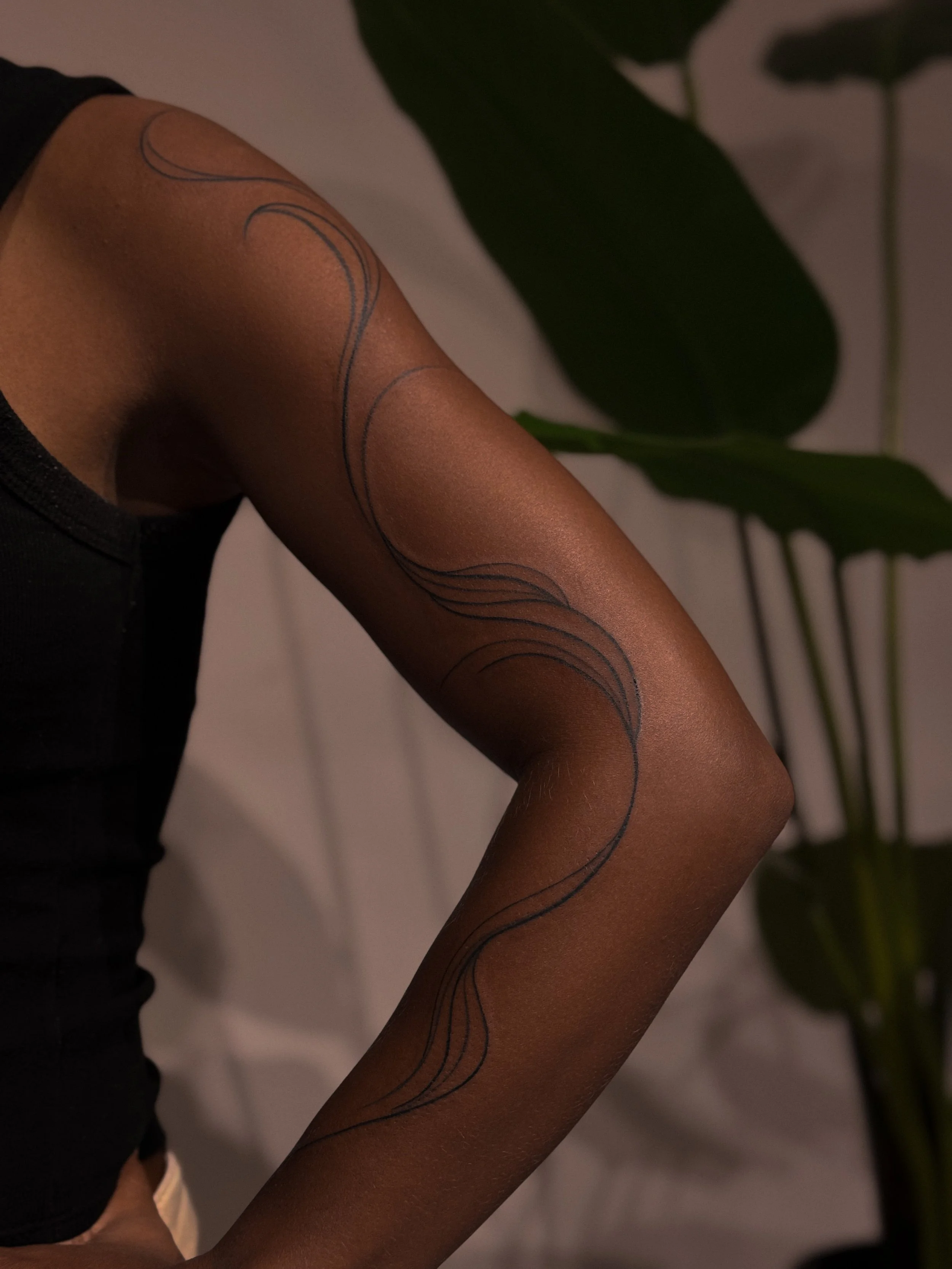 Close-up of a person's arm wearing sheer black stockings with a black ink line drawing tattoo of flowing, curved lines, with a plant with large green leaves in the background.