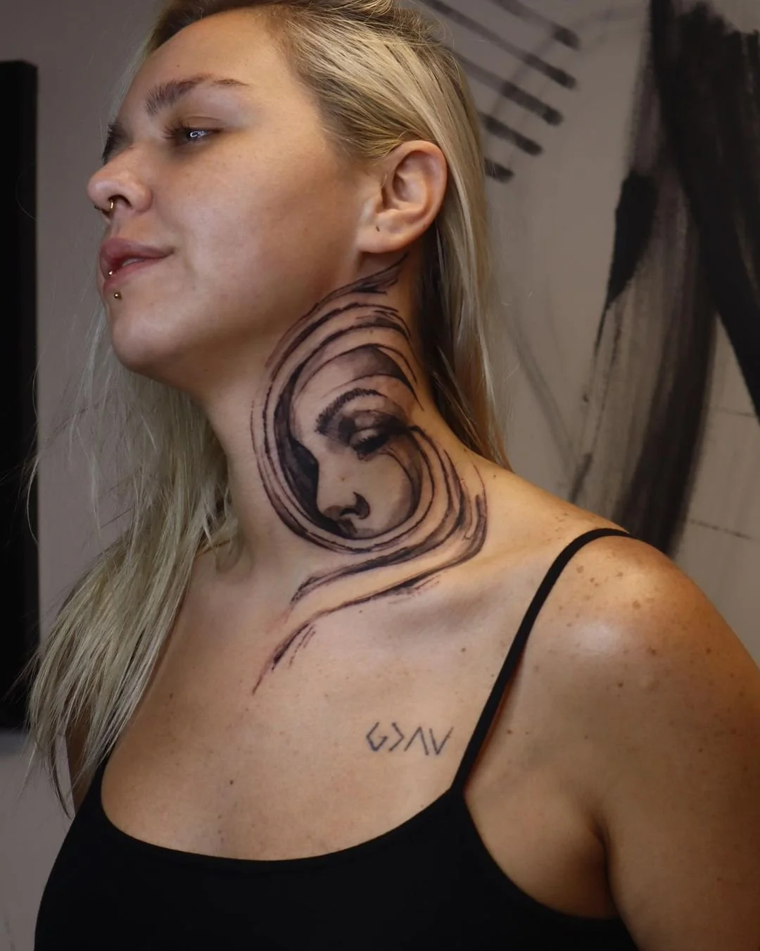 Person with abstract face tattoo on neck and small geometric tattoo on shoulder, wearing a black top.