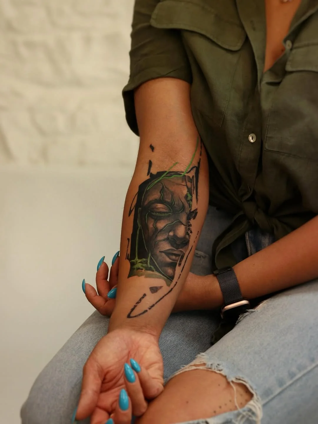 Tattoo of a woman's face with closed eyes on a person's forearm, with black and green ink, next to ripped jeans and a green shirt.