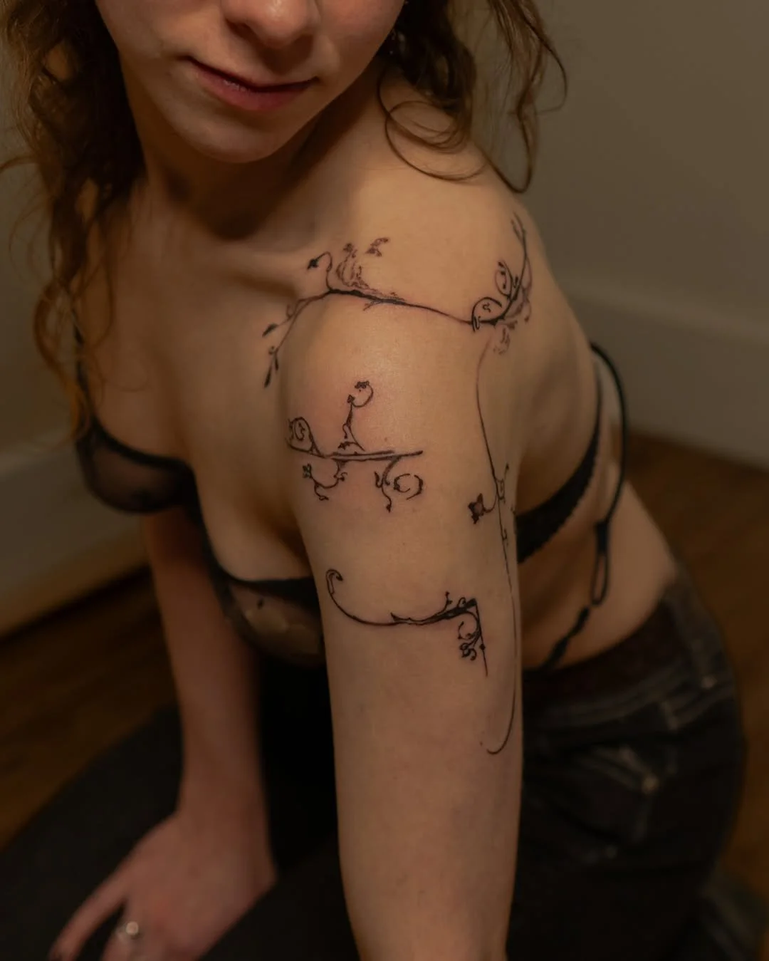 Woman with intricate linework tattoo on shoulder and arm, wearing a dark bra, sitting indoors.
