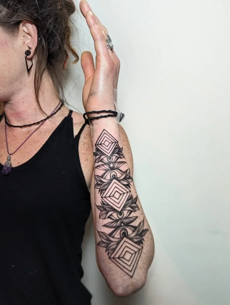 Close-up of a woman with tattoos on her arm, wearing a black sleeveless top, jewelry, and making a peace sign gesture with her hand against a plain background.