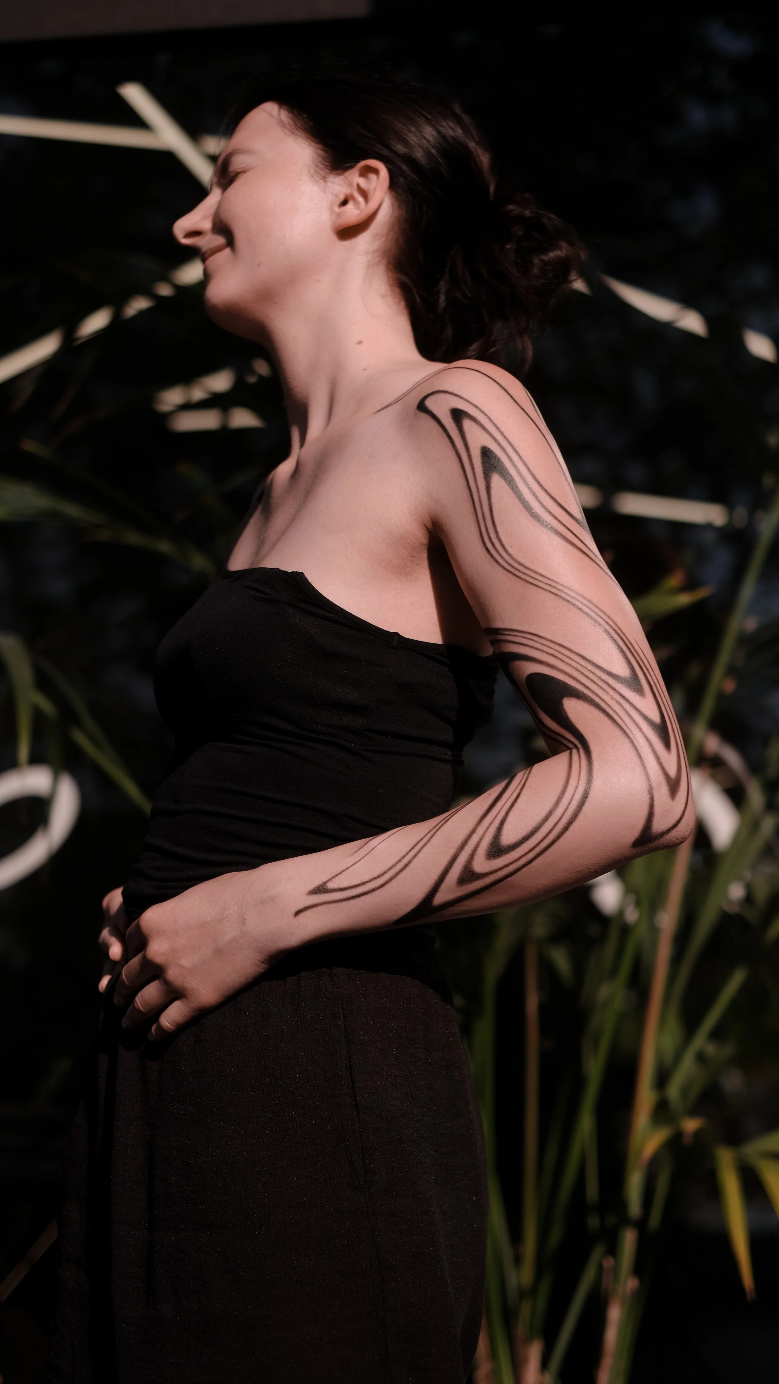 Person with large abstract black tattoo on arm, wearing black top, standing outdoors with foliage in background.