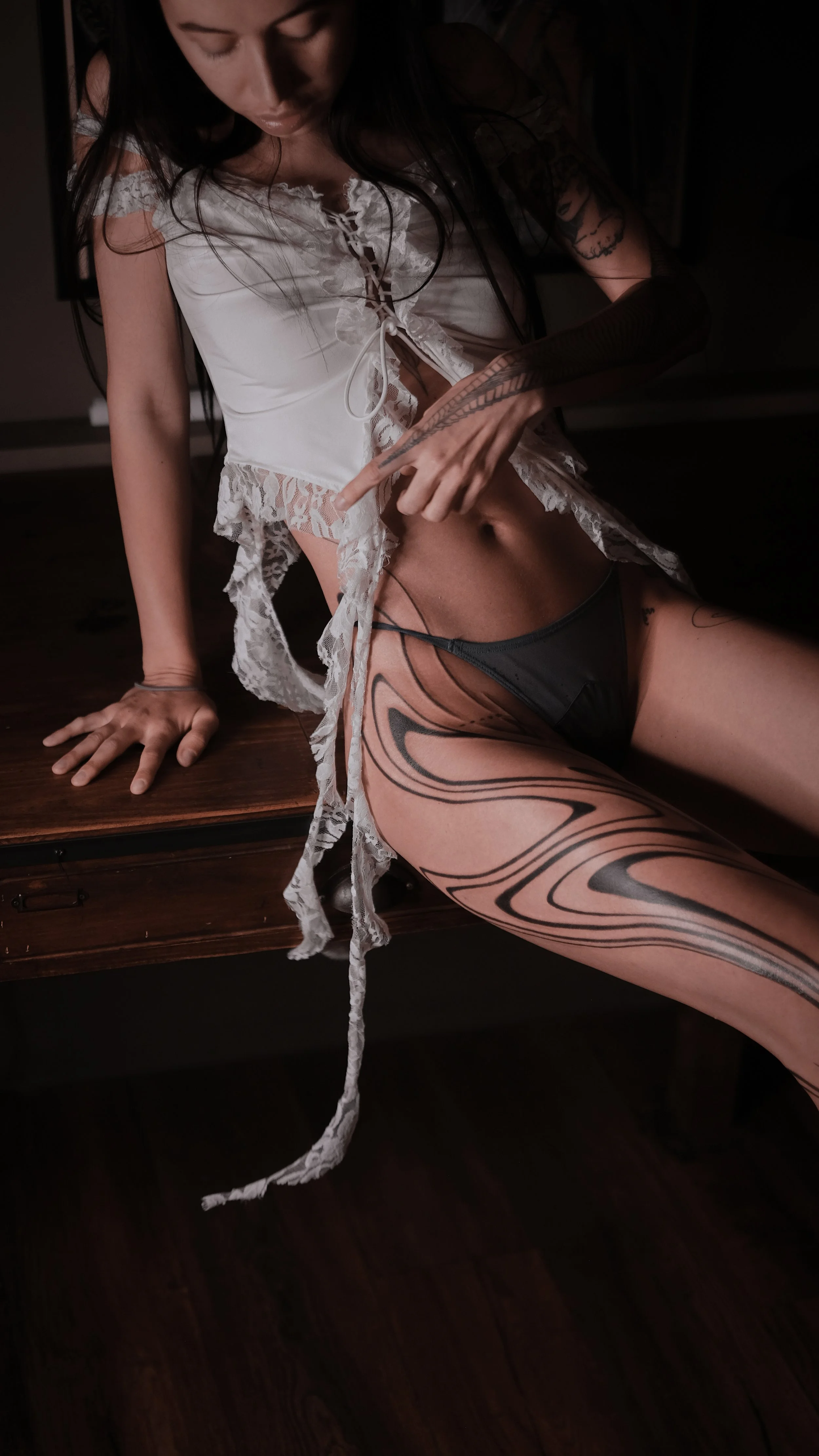 Person in white lace top and black underwear with leg tattoos sitting on a wooden surface.