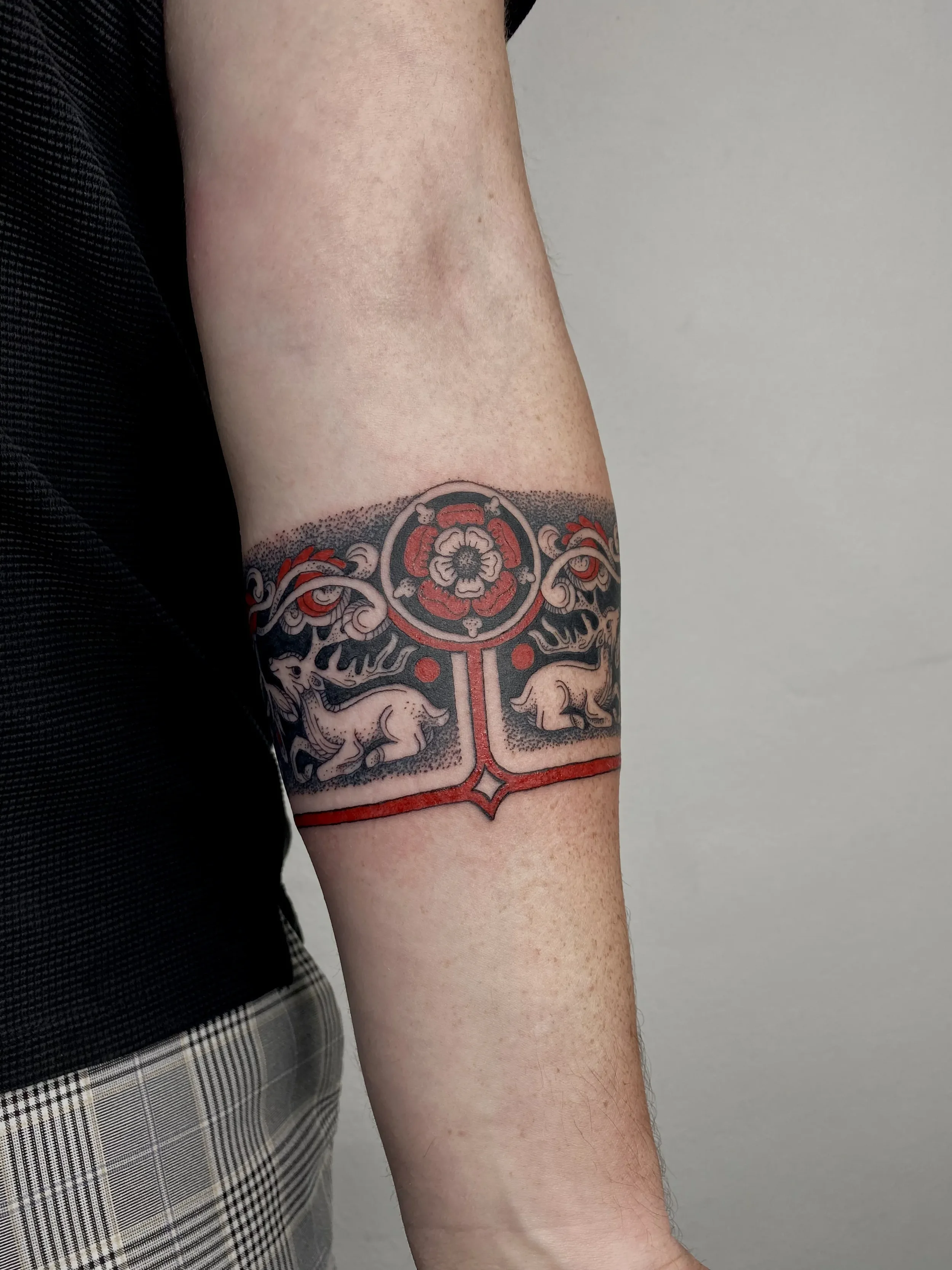Tattoo on person's upper arm featuring a circular floral design at the top, two animals resembling boars or pigs below, connected by a red geometric pattern.