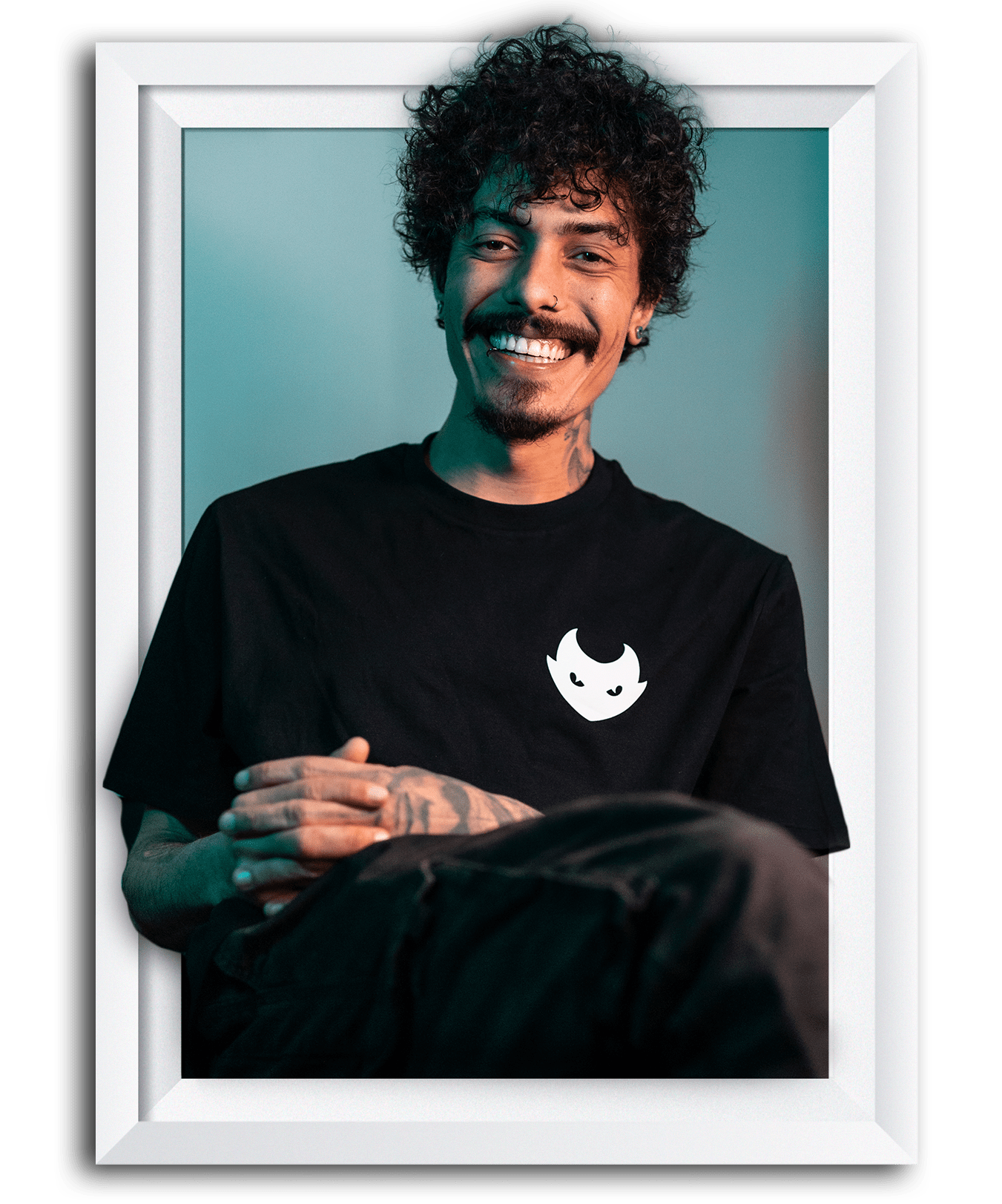 A smiling man with curly hair, tattoos, and a nose ring, wearing a black T-shirt with a white cartoon face logo, sitting against a teal background with a white frame around the image.
