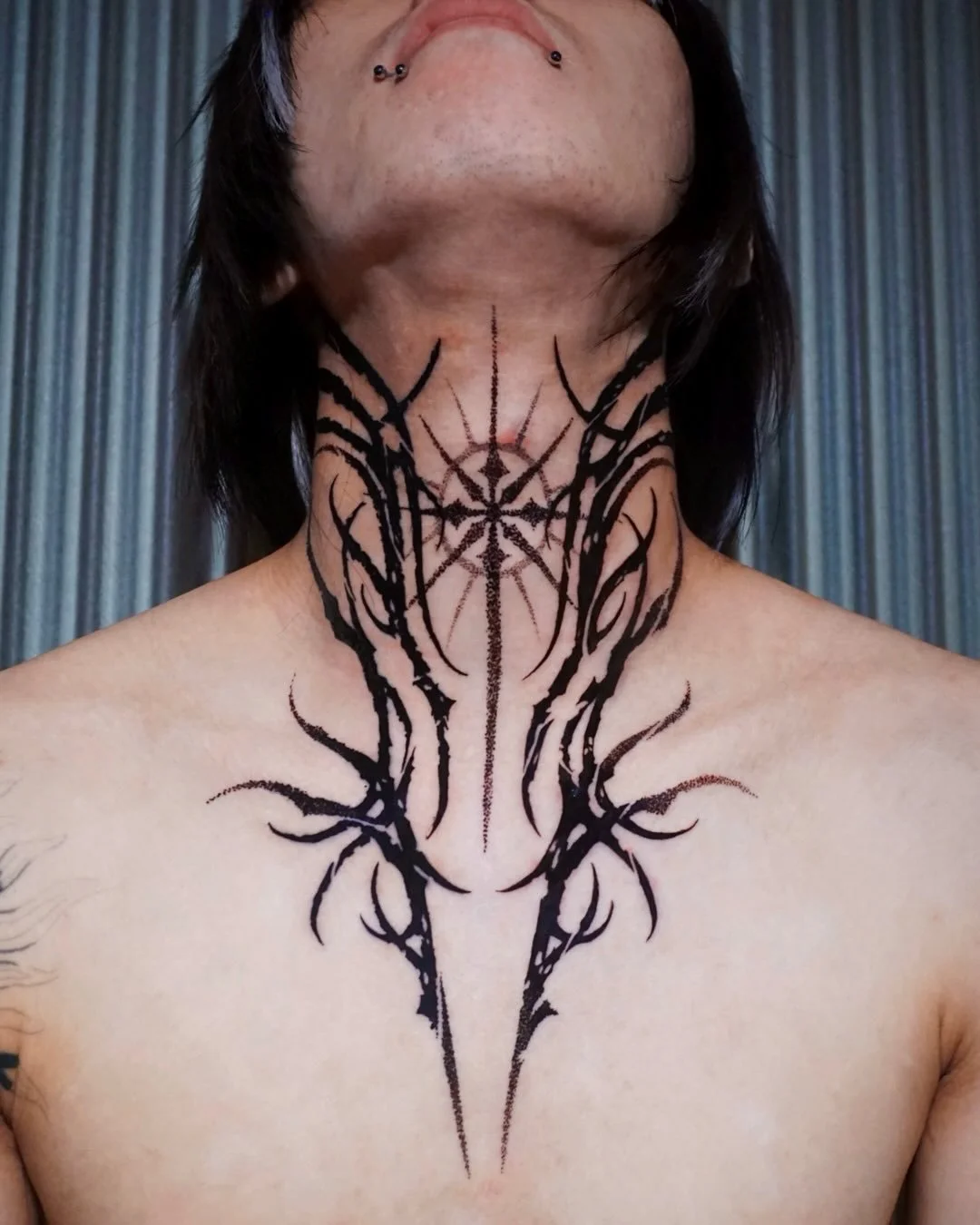 Person with black body art tattooed on their neck and chest, featuring intricate abstract designs and symmetrical patterns.