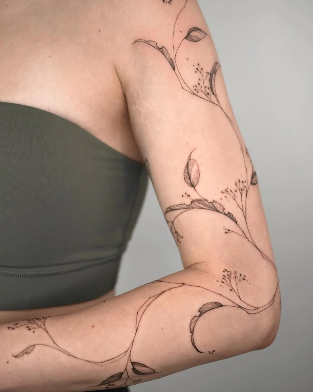 Tattoo of delicate botanical line art with leaves and small flowers on a person's arm and shoulder.