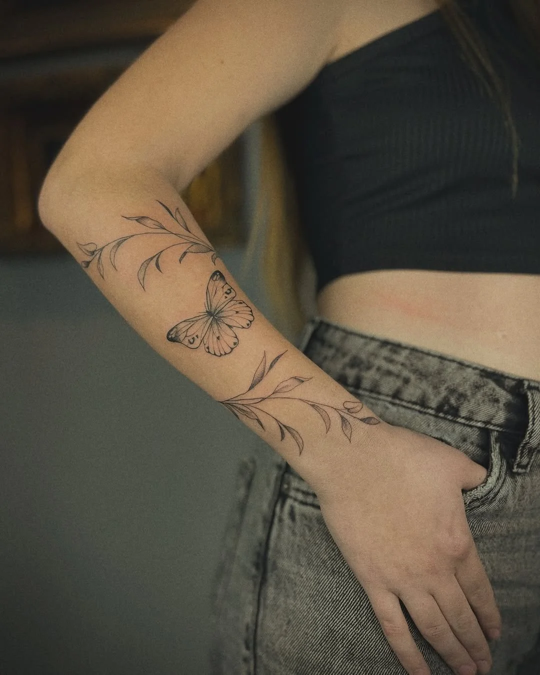 A person's arm with a delicate tattoo of a butterfly and leafy branches, dressed in a black top and gray jeans.