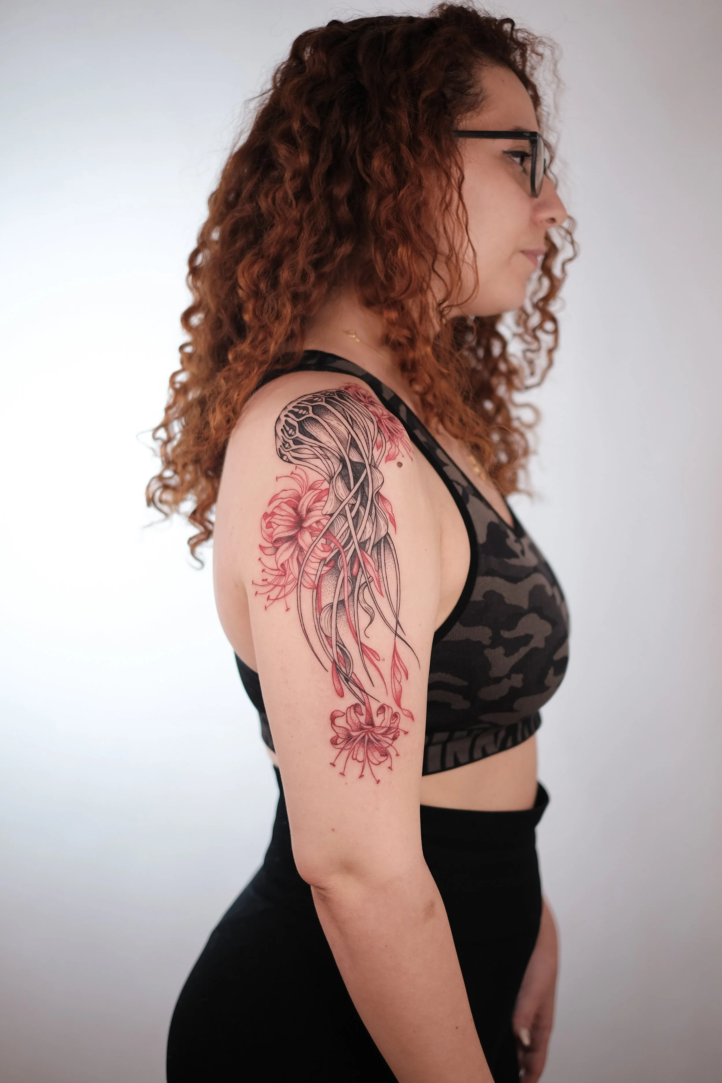 A woman with curly red hair, black glasses, wearing a black camouflage sports bra and black pants, standing sideways to the camera, showing a large tattoo of floral and abstract designs on her upper arm.