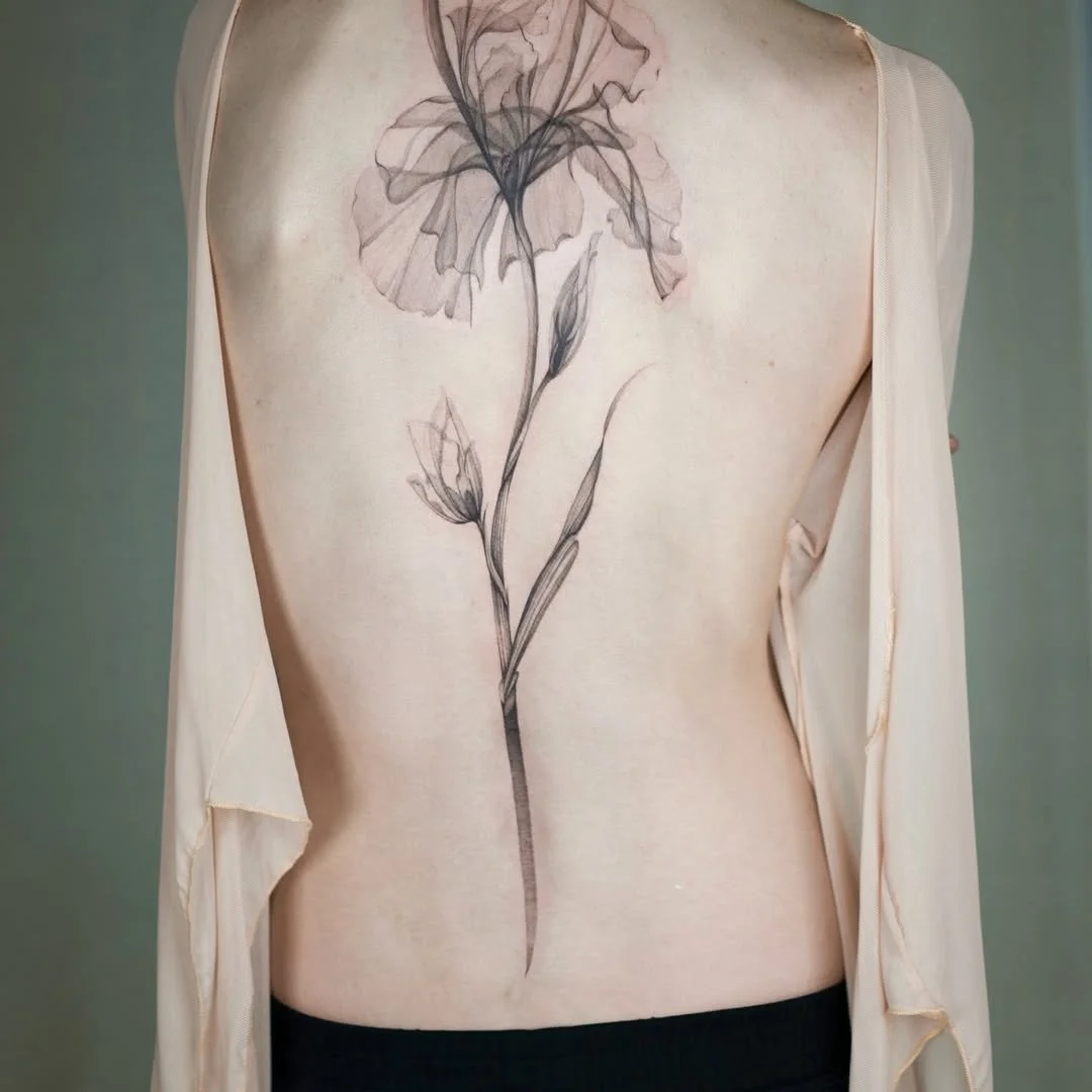 A person's back with a large, detailed black ink tattoo of a flower extending from the shoulder blade down to the waist.