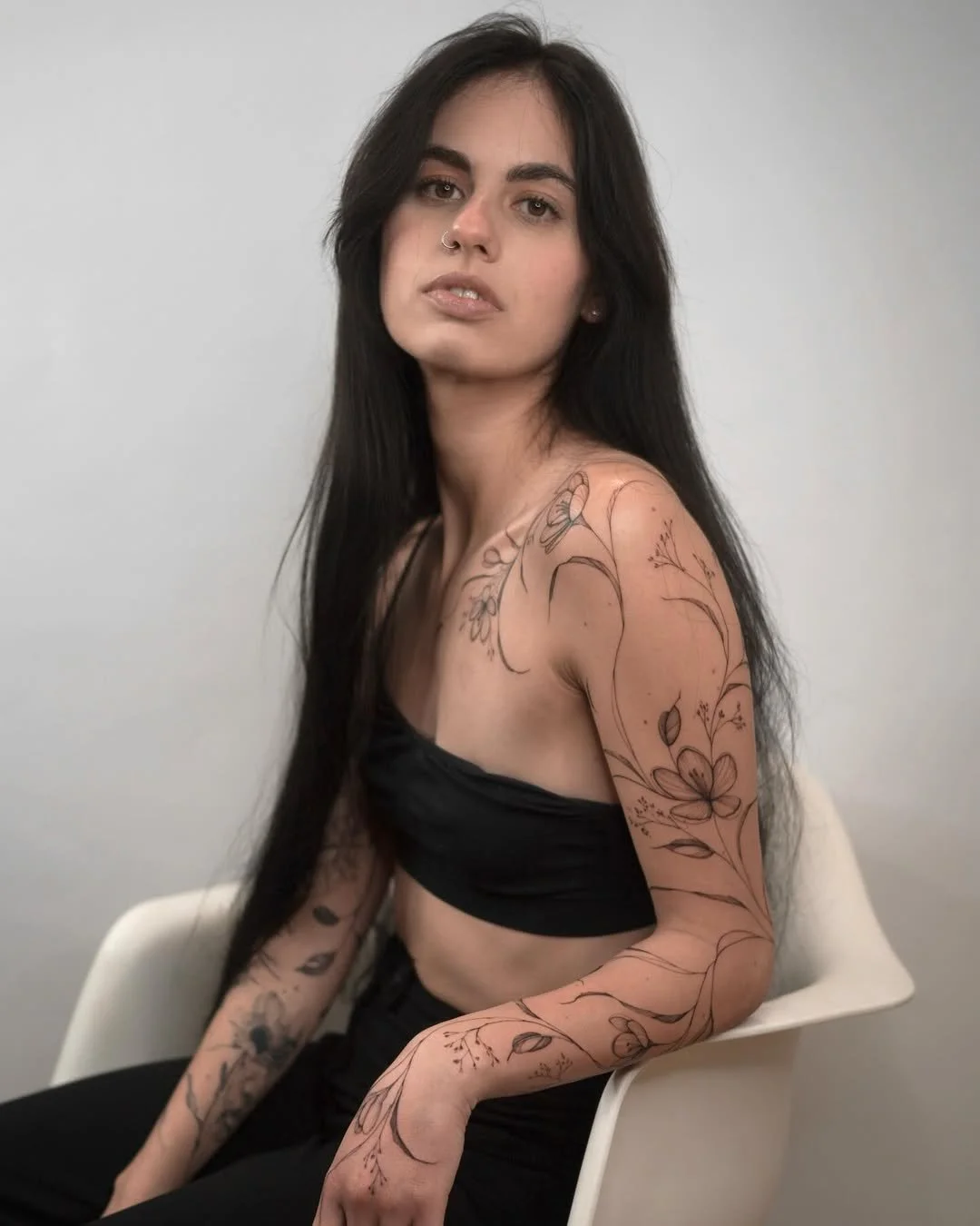 Young woman with long black hair, wearing a black strapless top and black pants, sitting on a white chair against a plain white wall. She has a nose piercing and visible arm tattoos with floral designs.