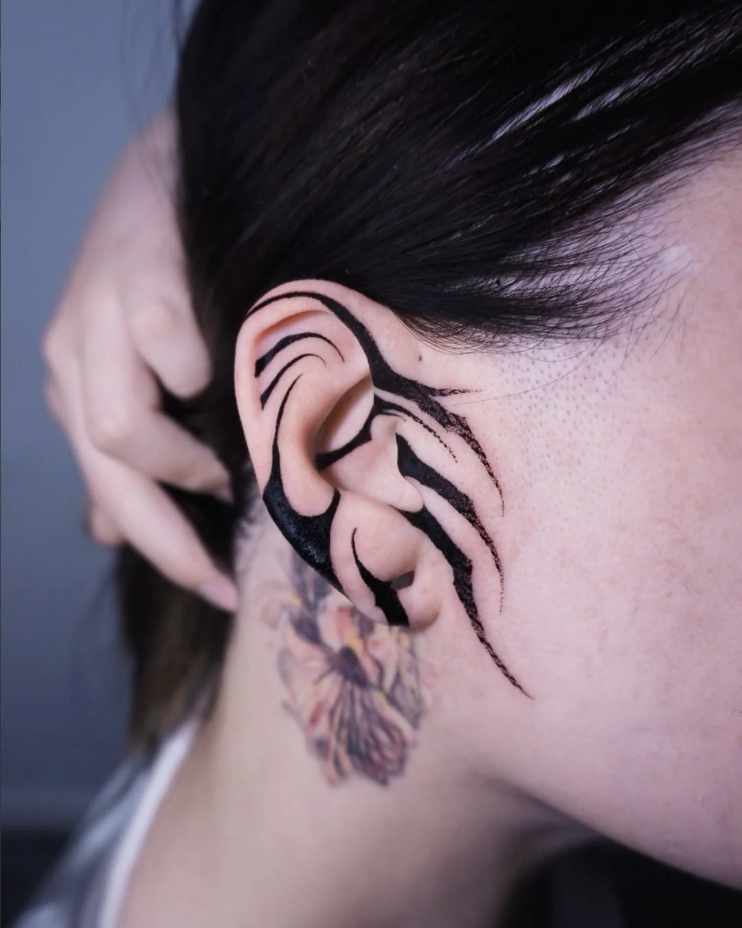 Close-up of a person's ear with black abstract tattoo art extending from the ear to the neck, dark hair, and part of a floral tattoo on the neck.