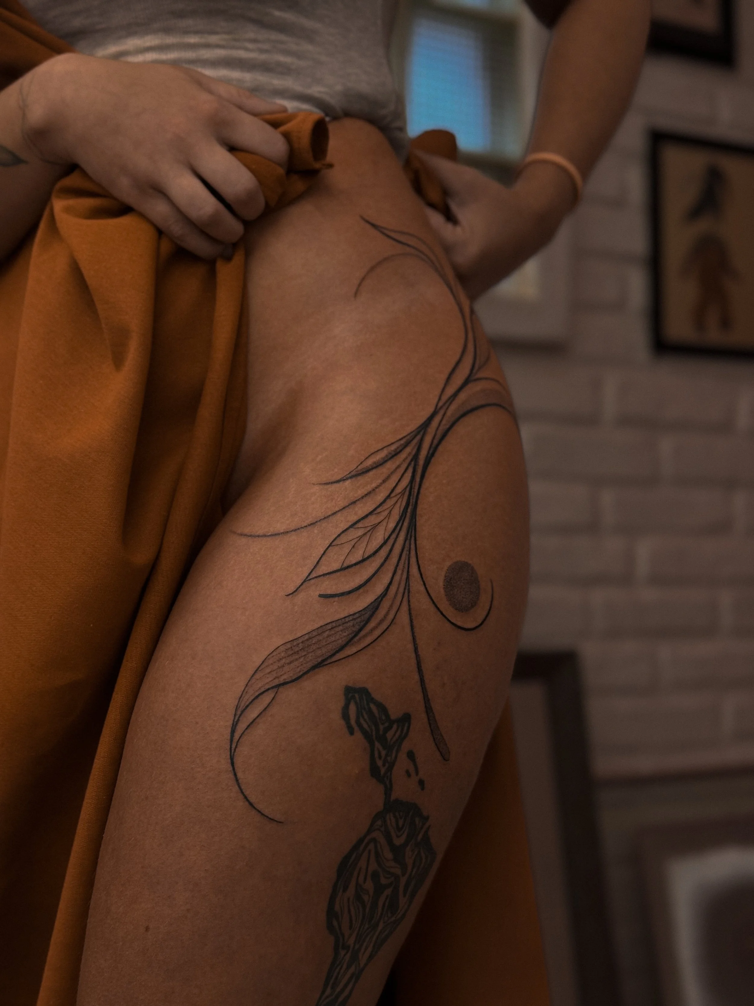 A person lifting a brown skirt reveals a thigh tattoo featuring a book with flowing lines and a circular shape.