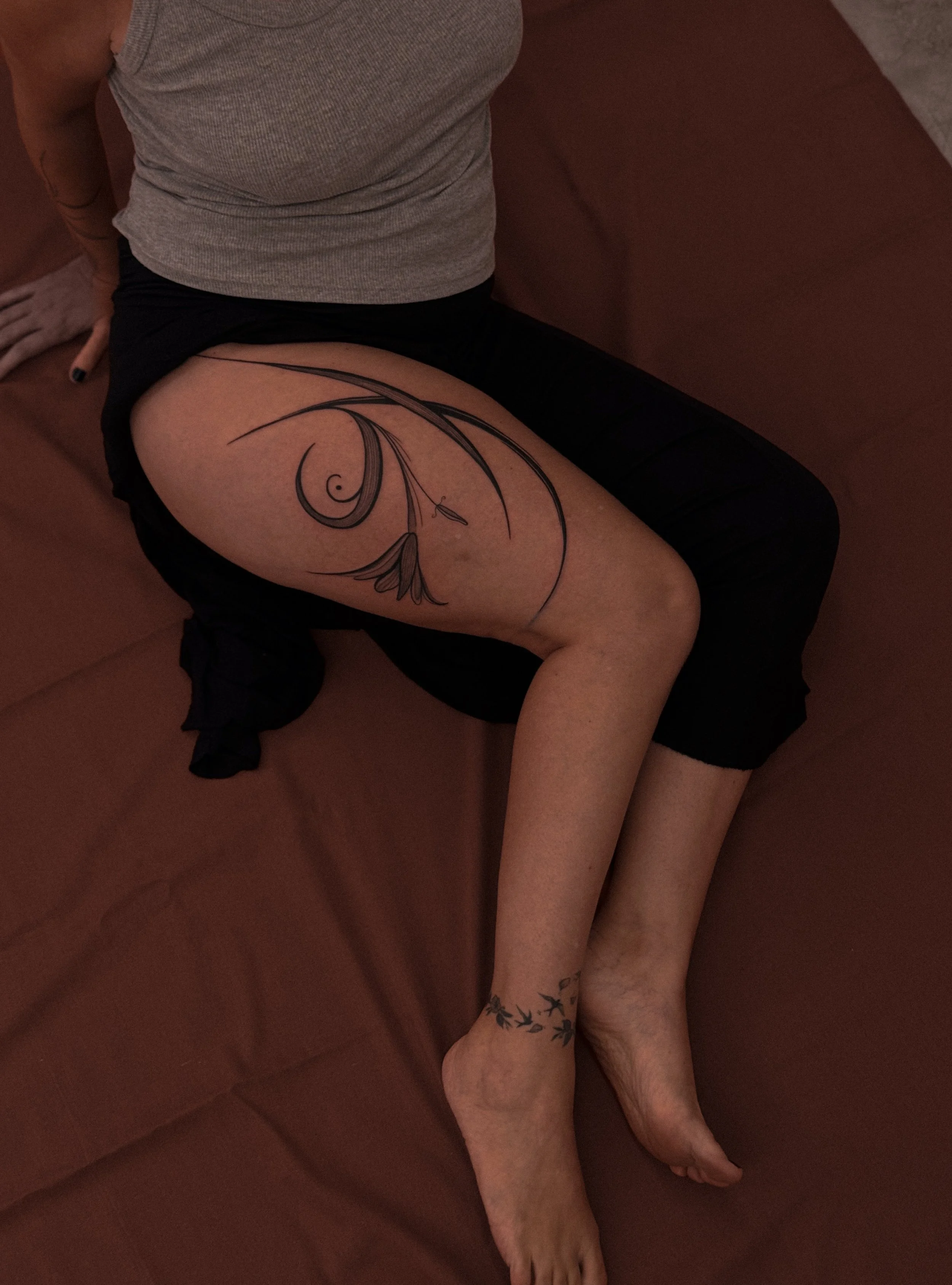 A woman sitting on a sofa, with a large abstract tattoo on her thigh and a smaller tattoo on her ankle.