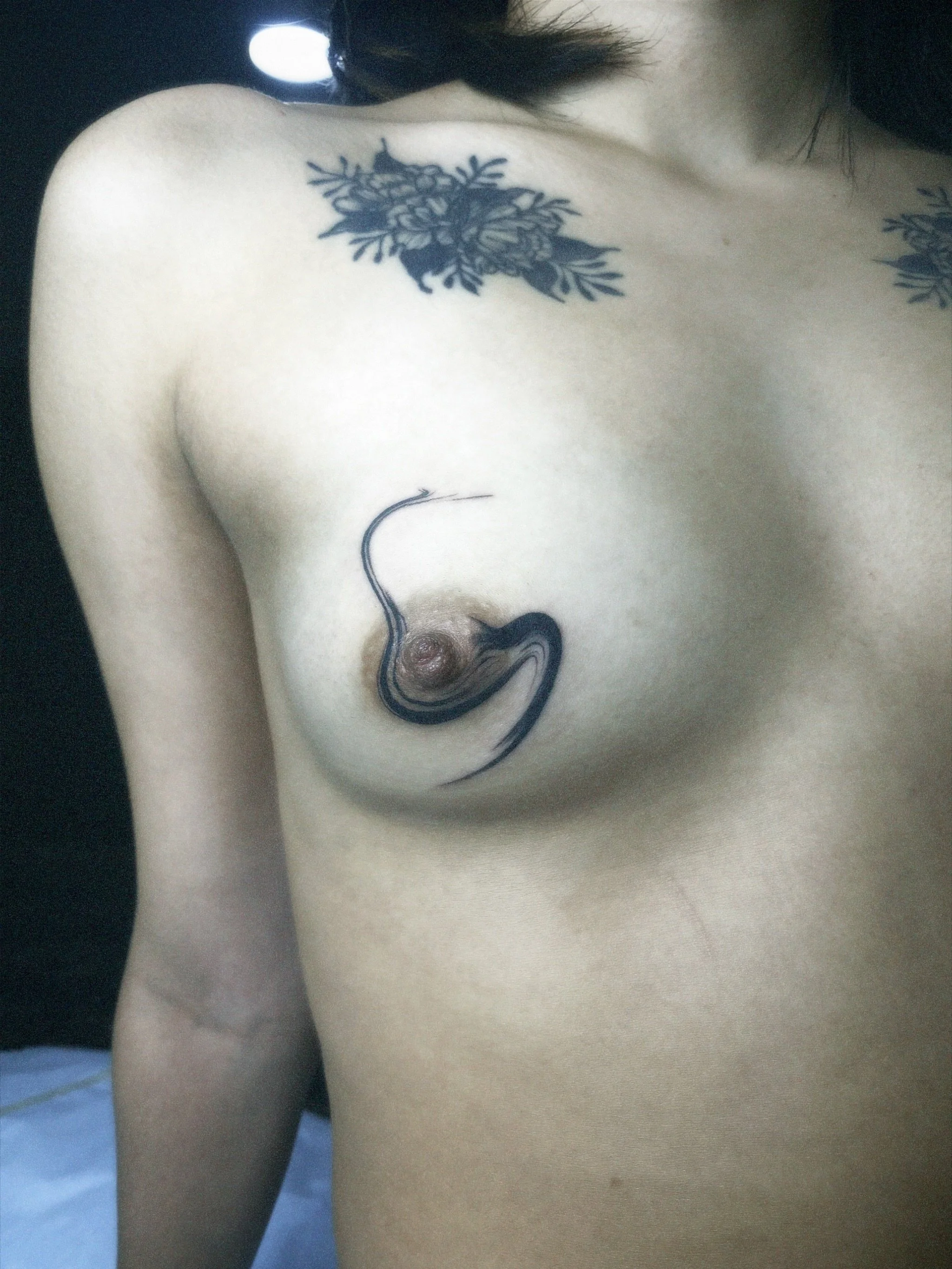 A person with a floral tattoo on their shoulder and inky black tattoo on their chest, featuring an eel-like creature weaving around their breast.