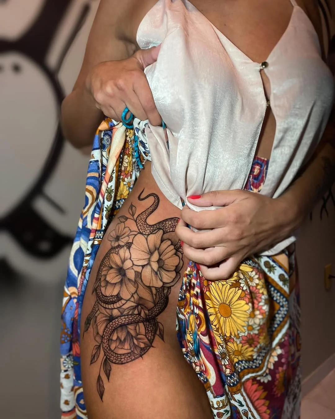 Person lifting their white satin top to reveal a tattoo of a snake entwined with flowers on their upper thigh.