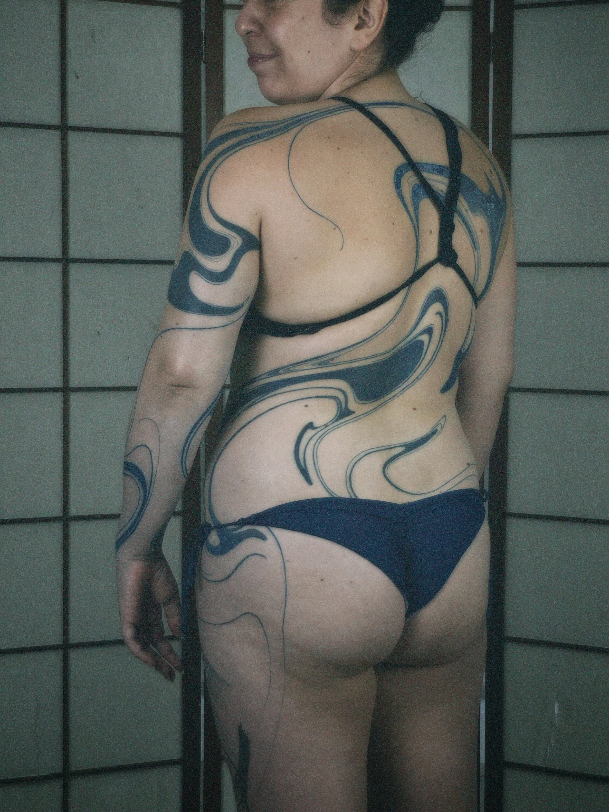 A woman with tattoos on her shoulder, arm, and back, wearing a black strappy bikini, standing in front of a Japanese-style room divider.