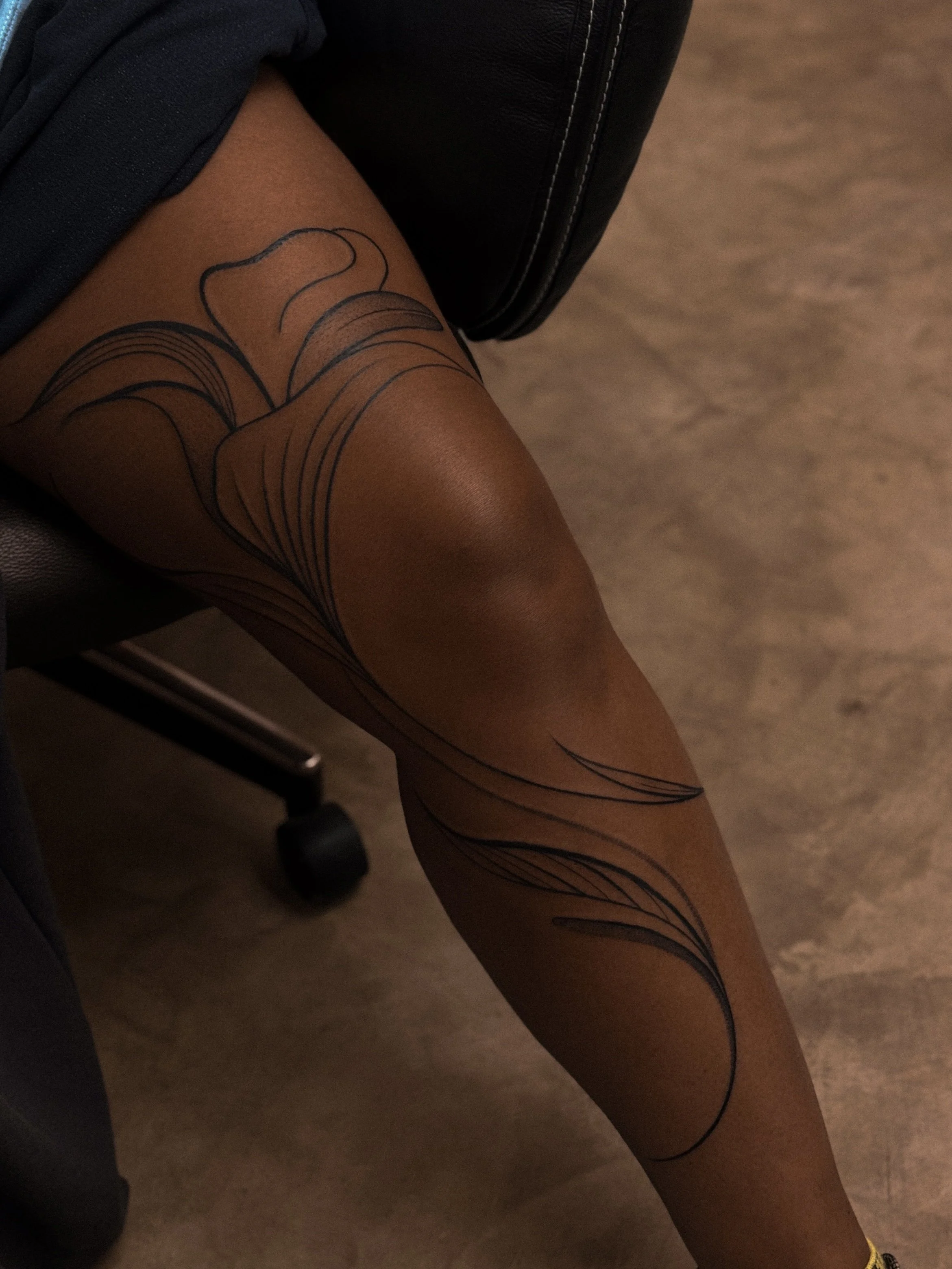 Close-up of a person's leg with black tattoo lines resembling abstract floral or leaf patterns, sitting on a chair.