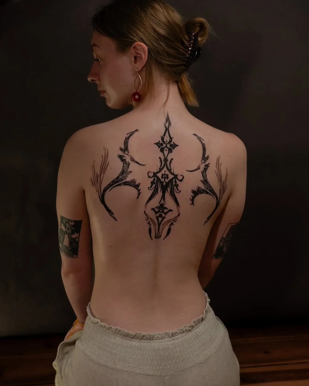 Person with intricate black back tattoo and earrings, facing away.