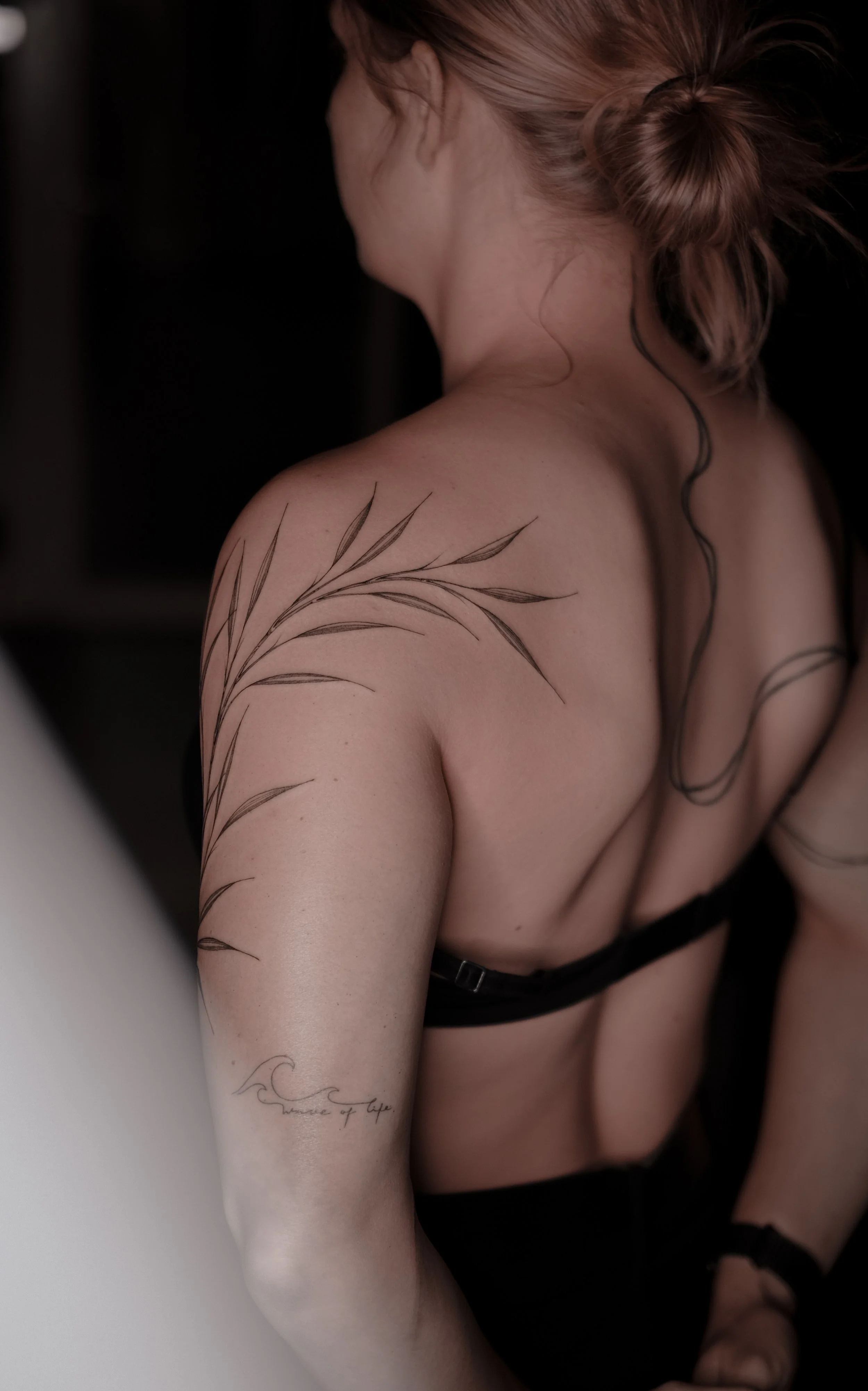 A person with long hair in a bun, wearing a black bra, showing tattoos on their back and arm.