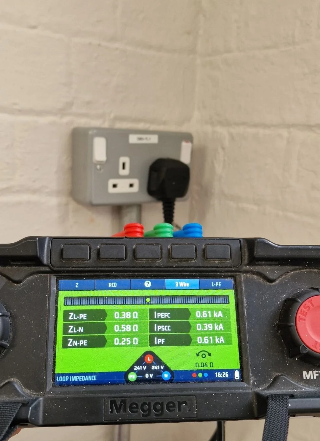 Multifunction tester showing loop impedance readings, plugged into a metal-clad socket on a painted brick wall