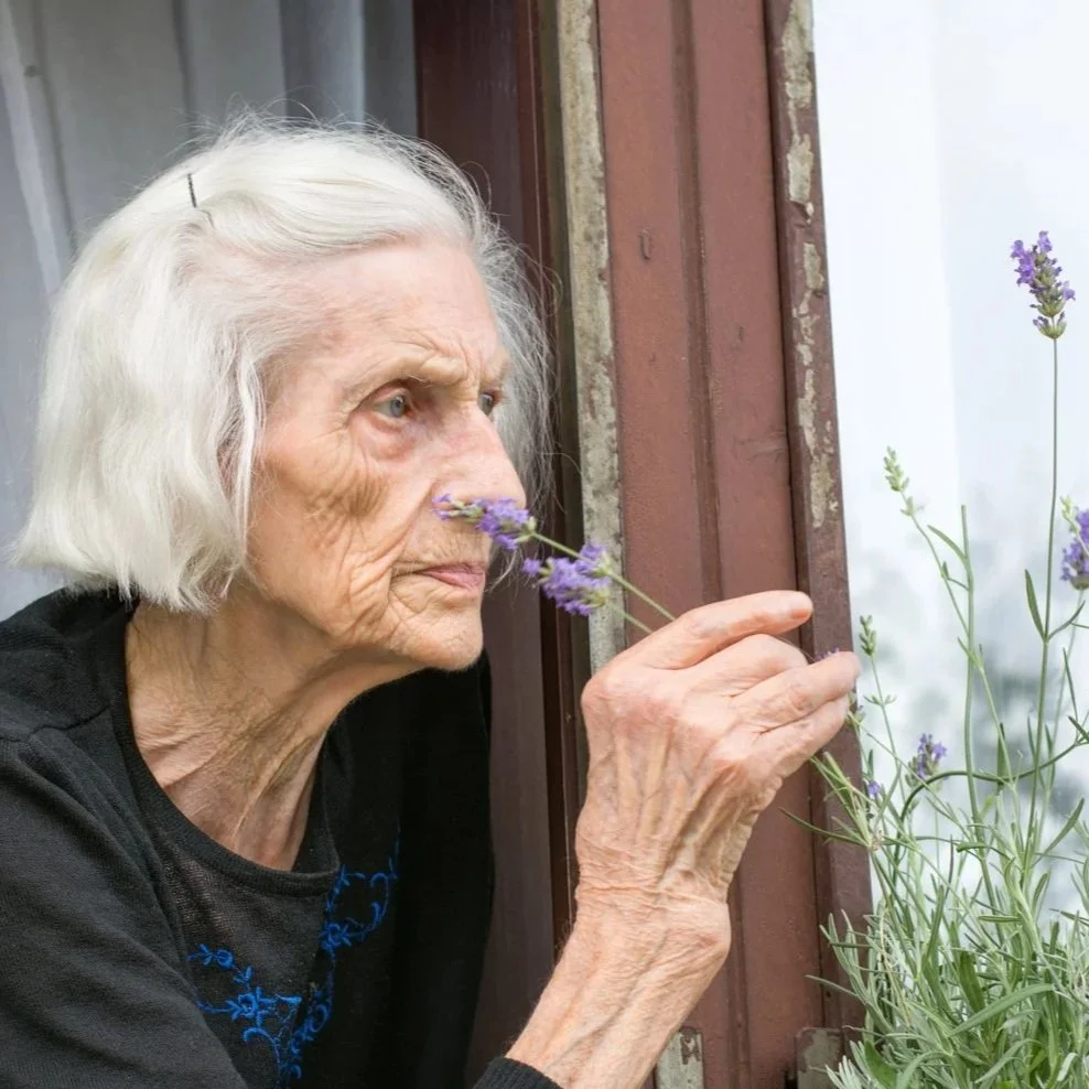A woman sitting on a white couch with her hands clasped, looking away with a contemplative expression, in a bright living room with white blinds and a small plant in the background.