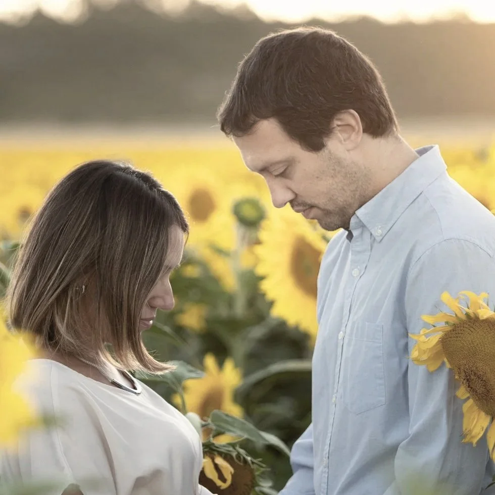 A man and woman embracing outdoors with trees in the background, the man kissing the woman's head as she closes her eyes.