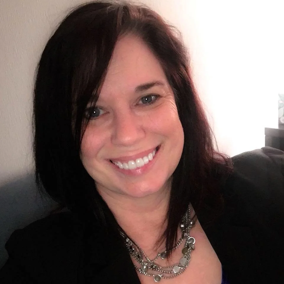 A smiling woman with shoulder-length dark hair and blue eyes, wearing a black top and layered silver necklaces, seated against a neutral background.