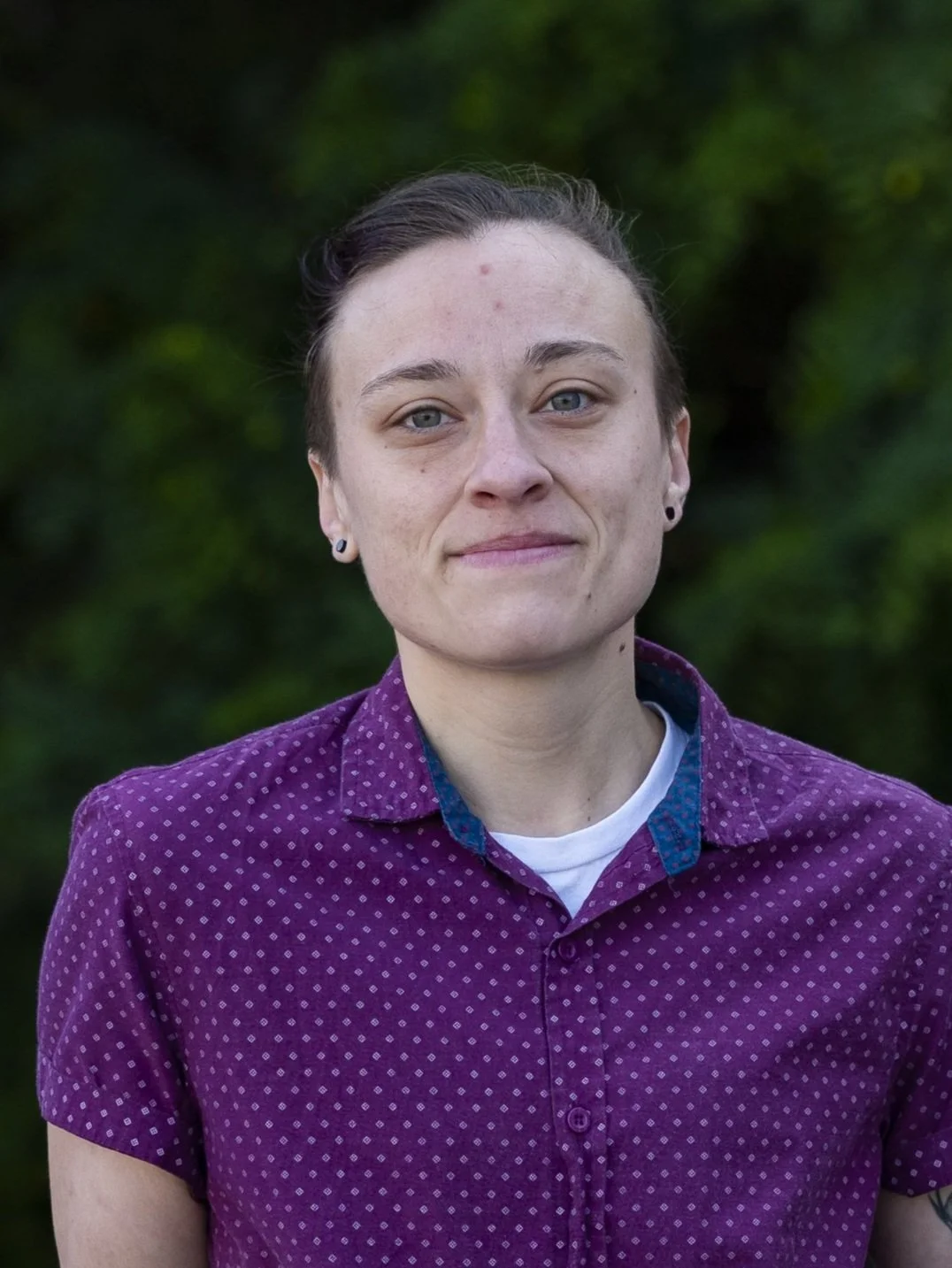 Person wearing a purple patterned shirt, standing outdoors against a green background.
