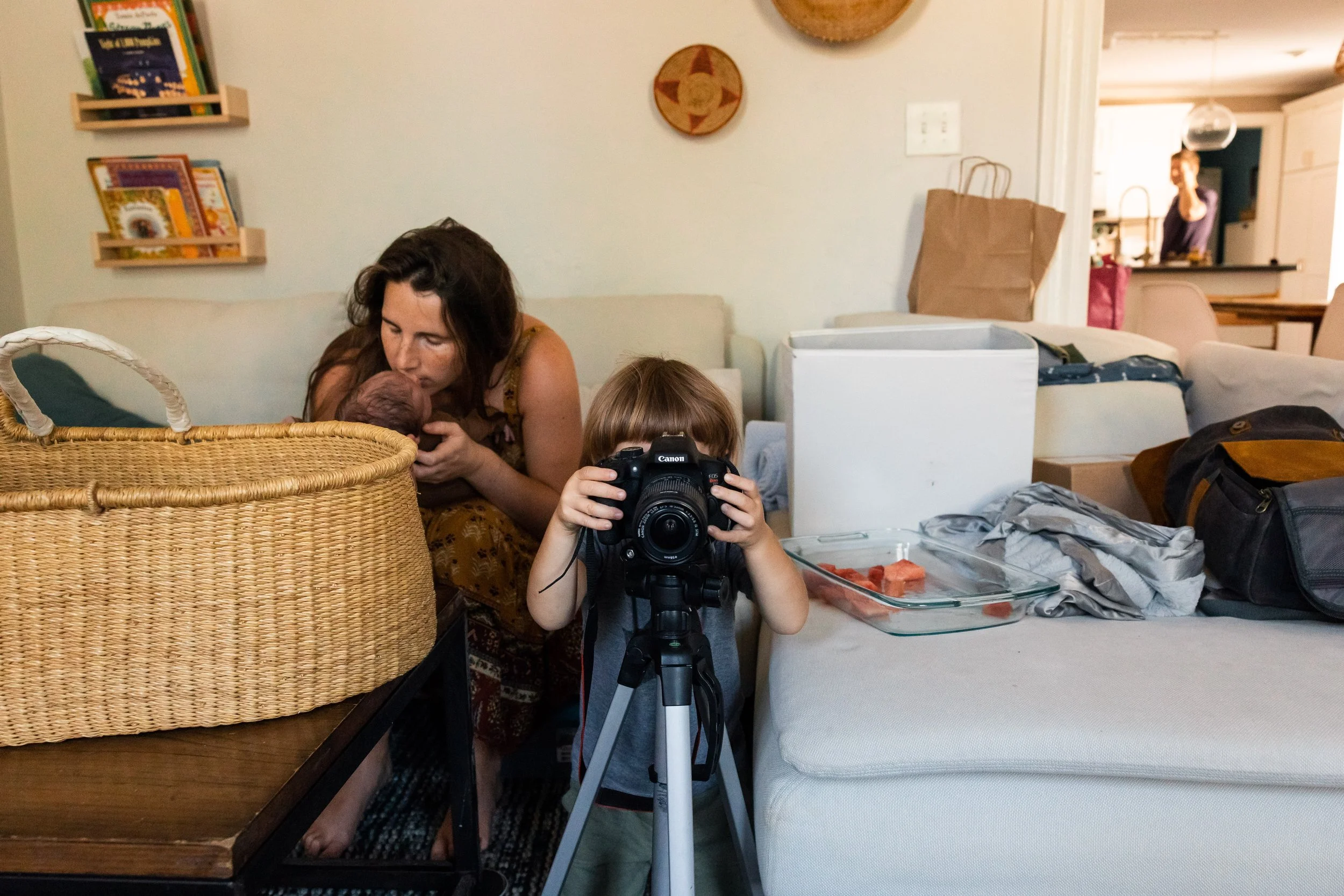 A woman is breastfeeding a newborn baby while a young boy with a camera on a tripod is taking a photo in a living room. The room has bookshelves, boxes, and a variety of household items.
