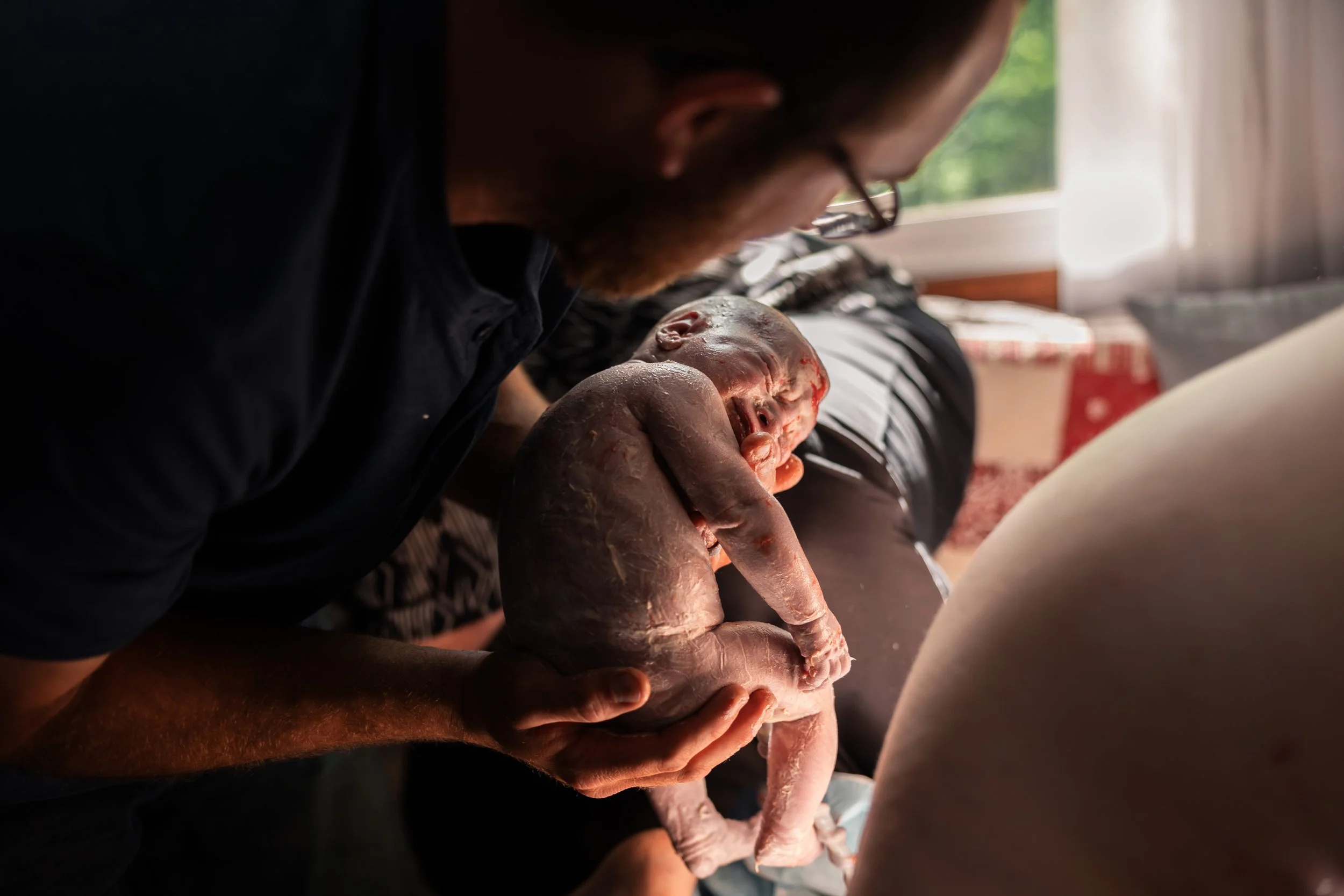 A man holding a newborn baby inside a home with natural light coming through the window.
