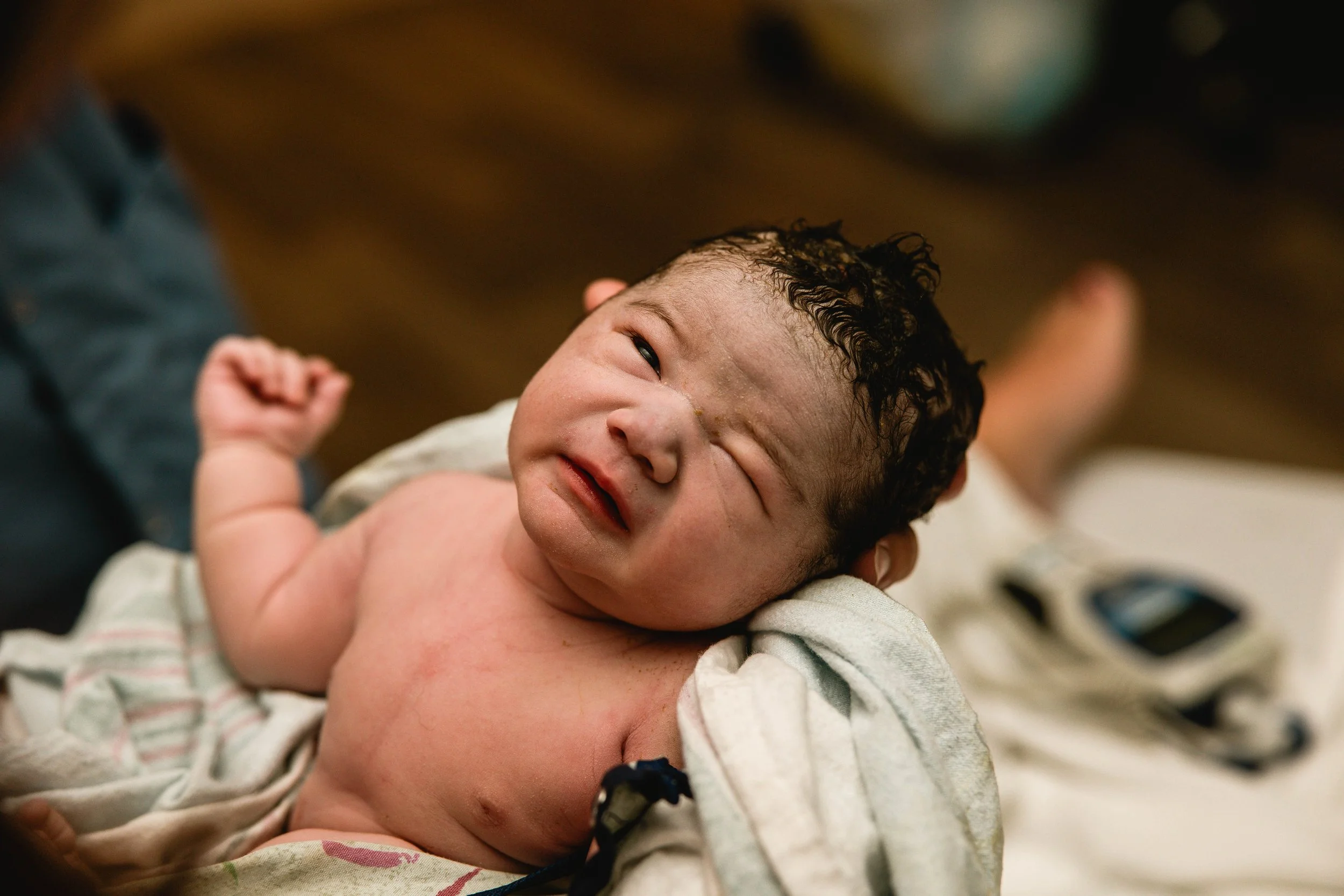 Close-up of a newborn baby lying on a hospital bed, with one eye partially closed, head turned to the side, and a neutral expression. The baby has dark, curly hair and is partially covered with a hospital blanket.
