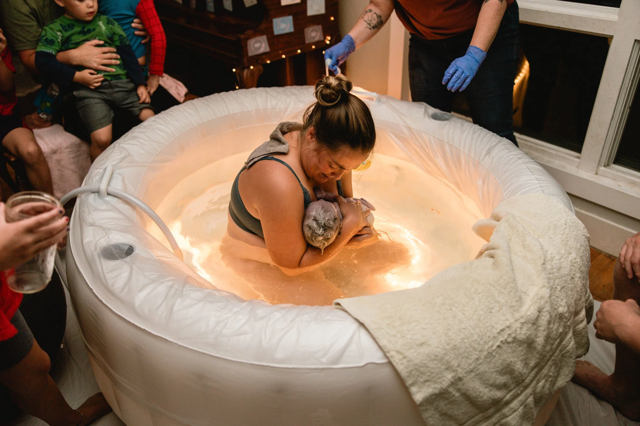 Woman holding a baby in a water birth tub during a water birth ceremony at a party surrounded by people.