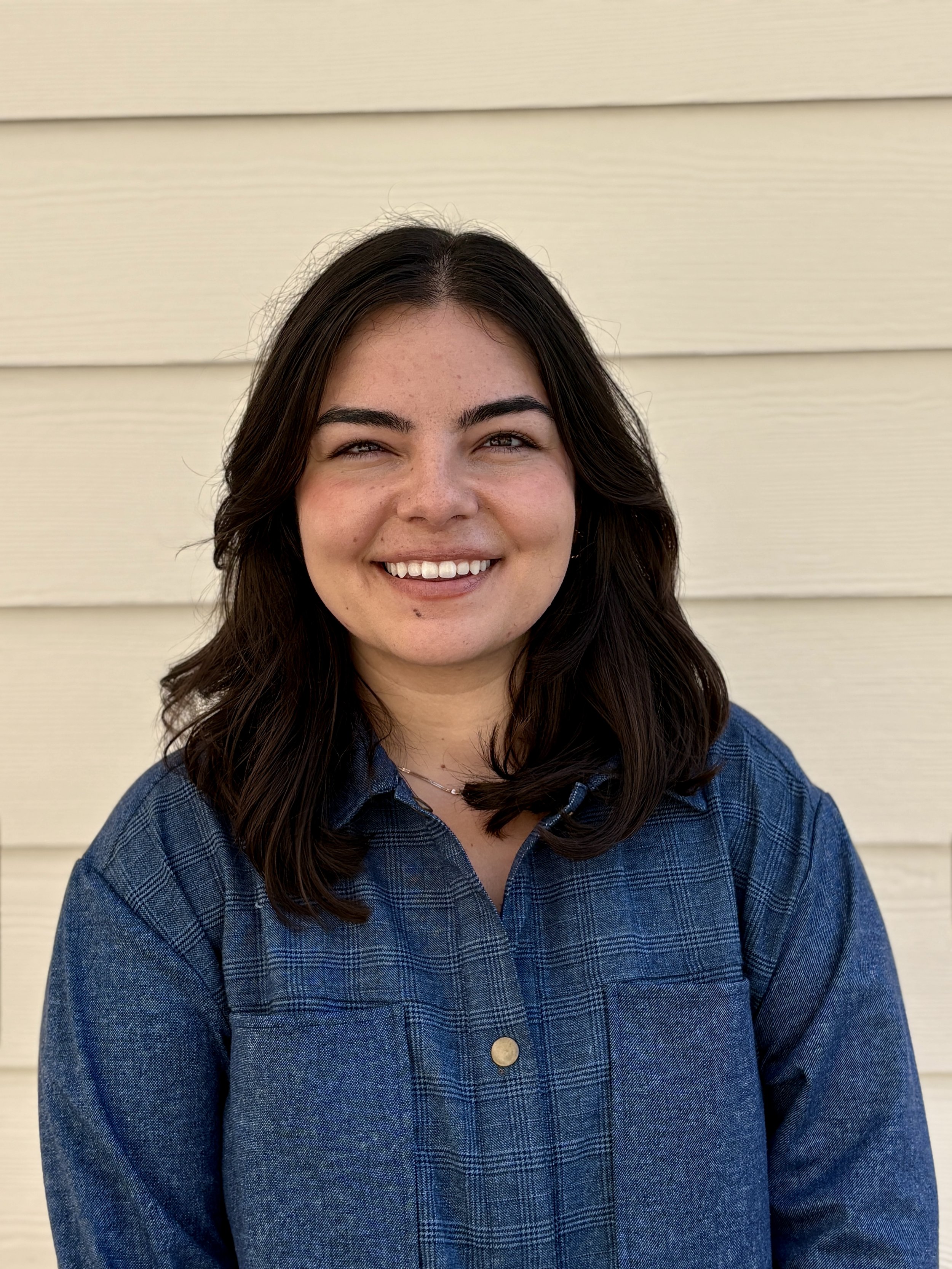 A young woman with dark brown hair, smiling, wearing a blue denim shirt, standing in front of a beige horizontal siding wall.