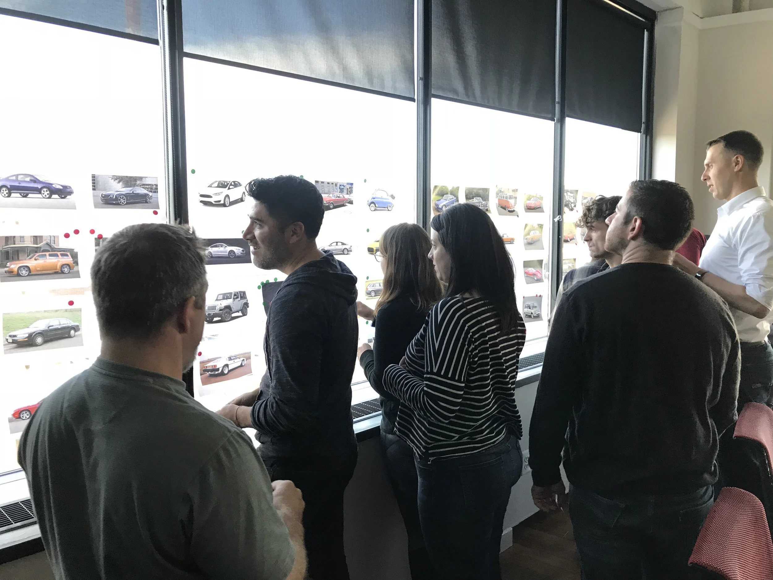 Group of people standing and sitting near large windows displaying photos of cars, with some individuals engaged in conversation.