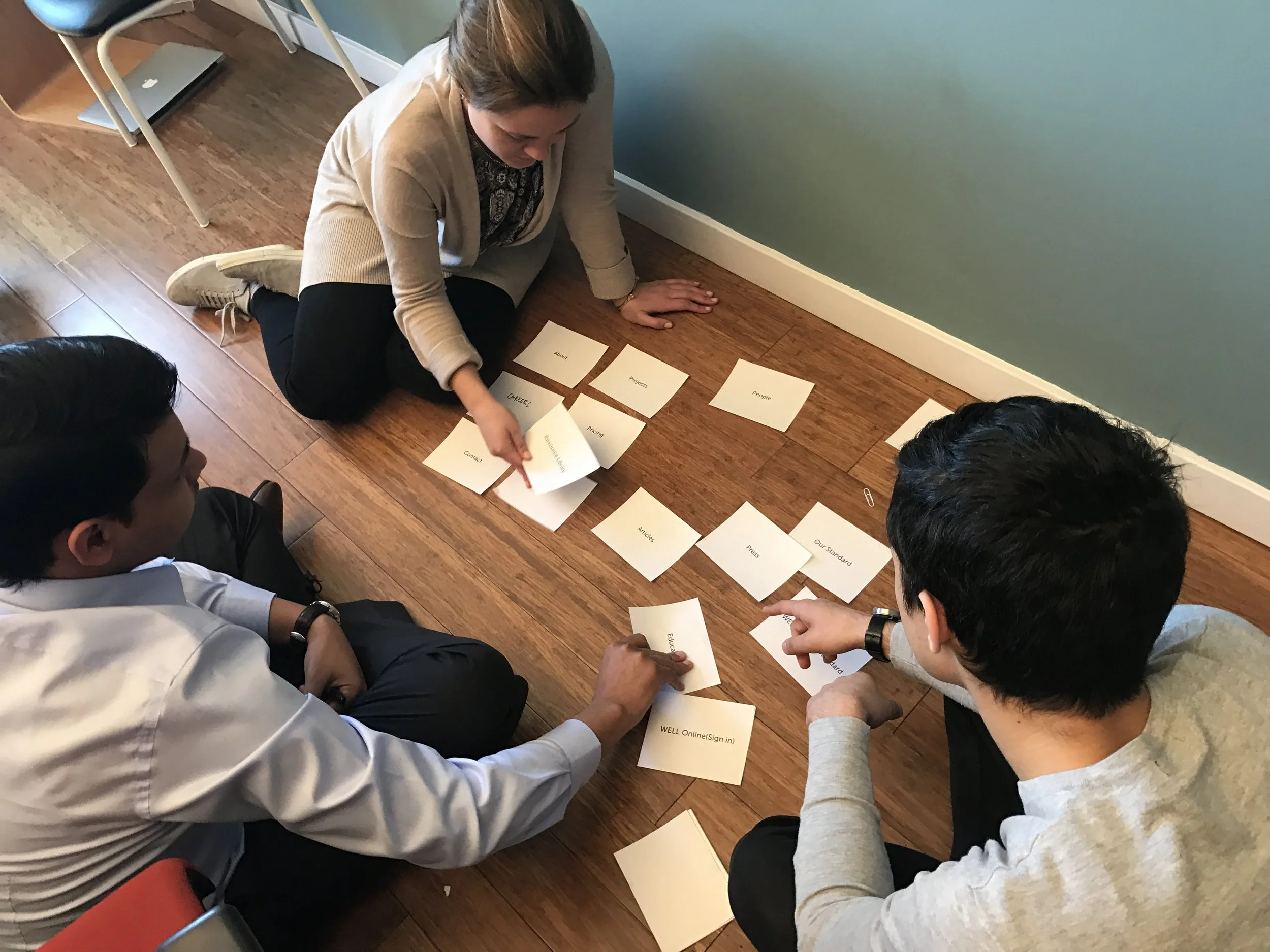 A group of people sitting on a wooden floor, engaging in a group activity involving arranging or reading printed cards with words such as 'Careers,' 'Projects,' and 'People.' A woman with short dark hair is pointing at one of the cards, and a man wea